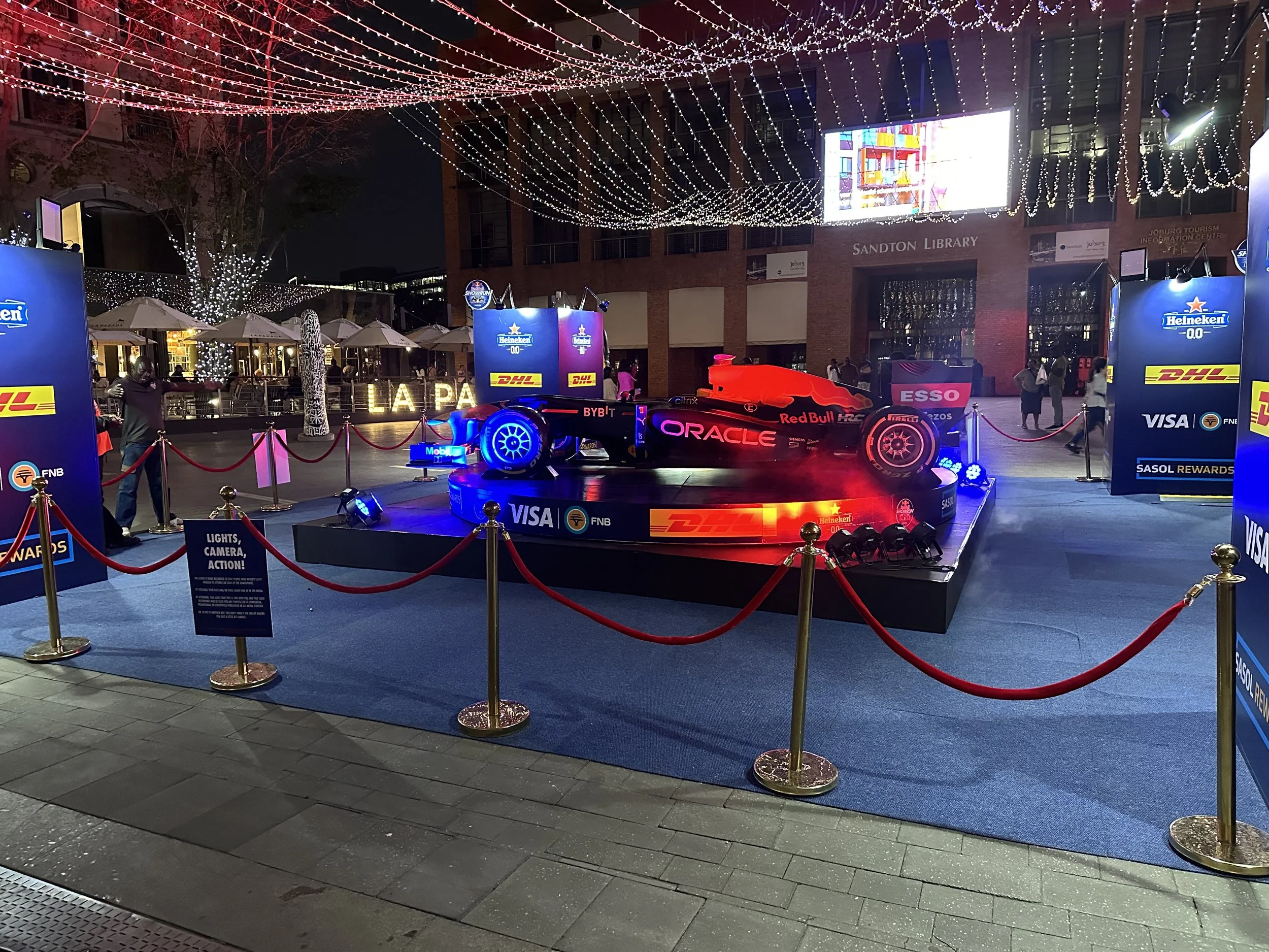 A Formula 1 race car on display at night, illuminated with bright lights and surrounded by a red rope barrier. The display area features sponsorship logos from Heineken, Visa, DHL, Oracle, Red Bull, and others, and is set outdoors with Christmas ligh