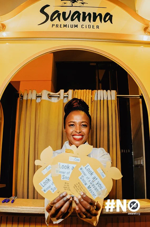 Woman smiling and holding promotional signs in front of a Savannah Premium Cider booth, with curtains behind her and the booth sign above.