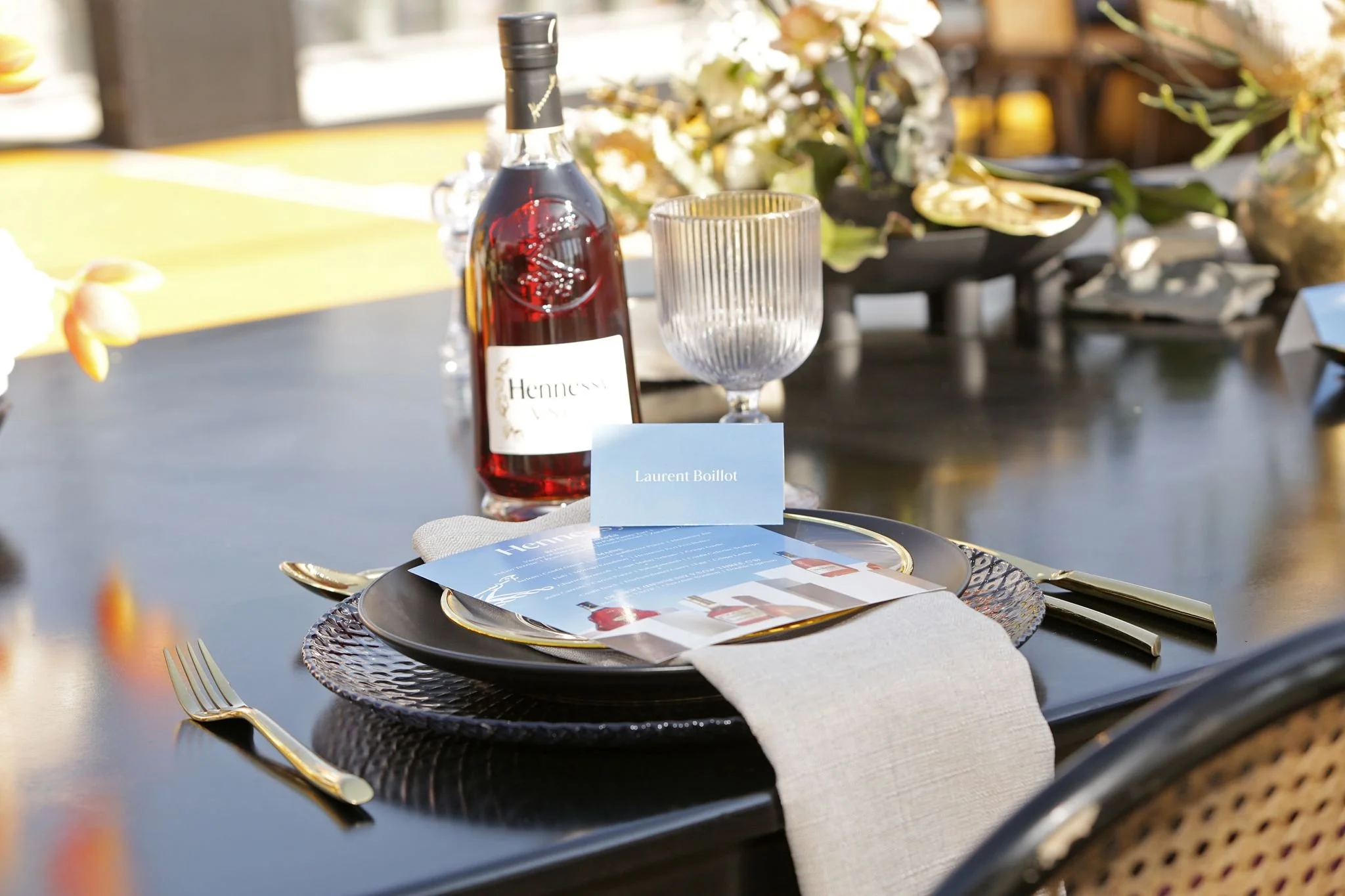 Elegant dining table with black plates, gold utensils, a glass, a bottle of Hennessy, and a place card reading Laurent Boillot, set near a window with natural sunlight.
