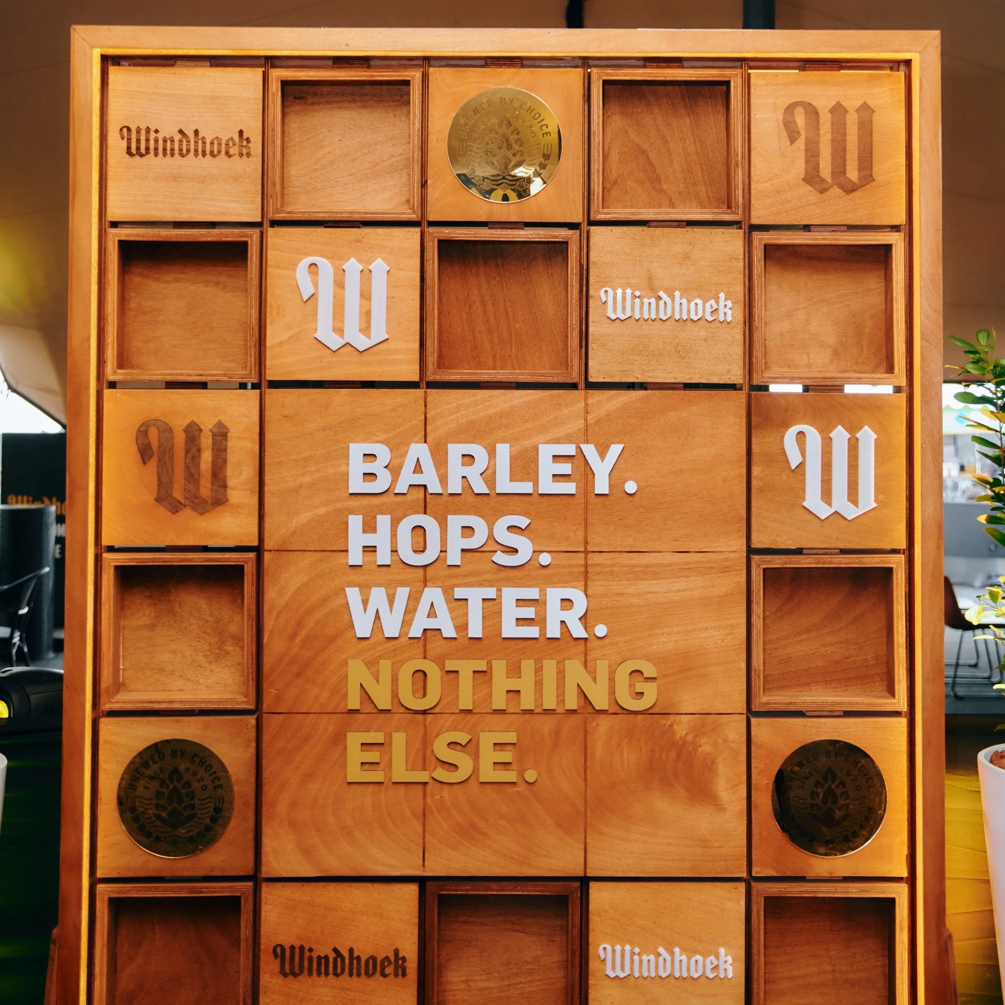A wooden display with compartments, featuring the words 'BARLEY. HOPS. WATER. NOTHING ELSE.' in white and gold letters, and the word 'Windhoek' written multiple times. There are also round black and gold labels.