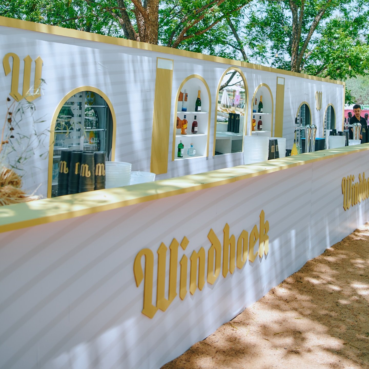 An outdoor bar with a white and gold design, displaying liquor bottles on shelves, and a man behind the bar, set among trees with green foliage.