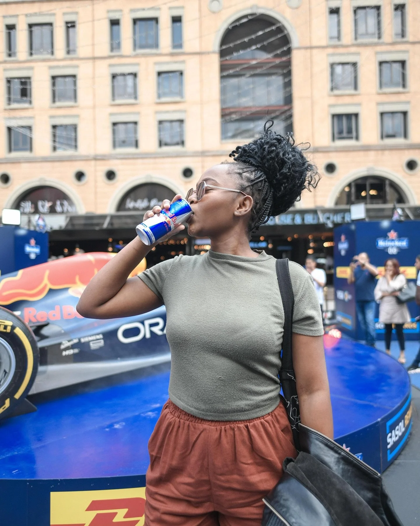 A woman with styled curly hair and sunglasses, wearing a green t-shirt and brown shorts, drinks Red Bull energy drink at a Formula 1 event, with a Red Bull Racing car in the background.