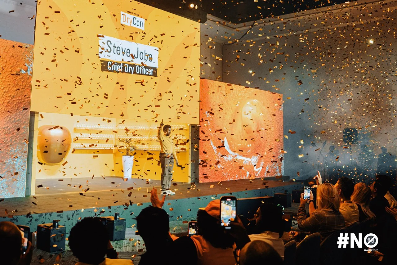 A person standing on stage at DryCon speaking to an audience, with confetti falling around, a large screen displaying his name and title, and audience members taking photos.