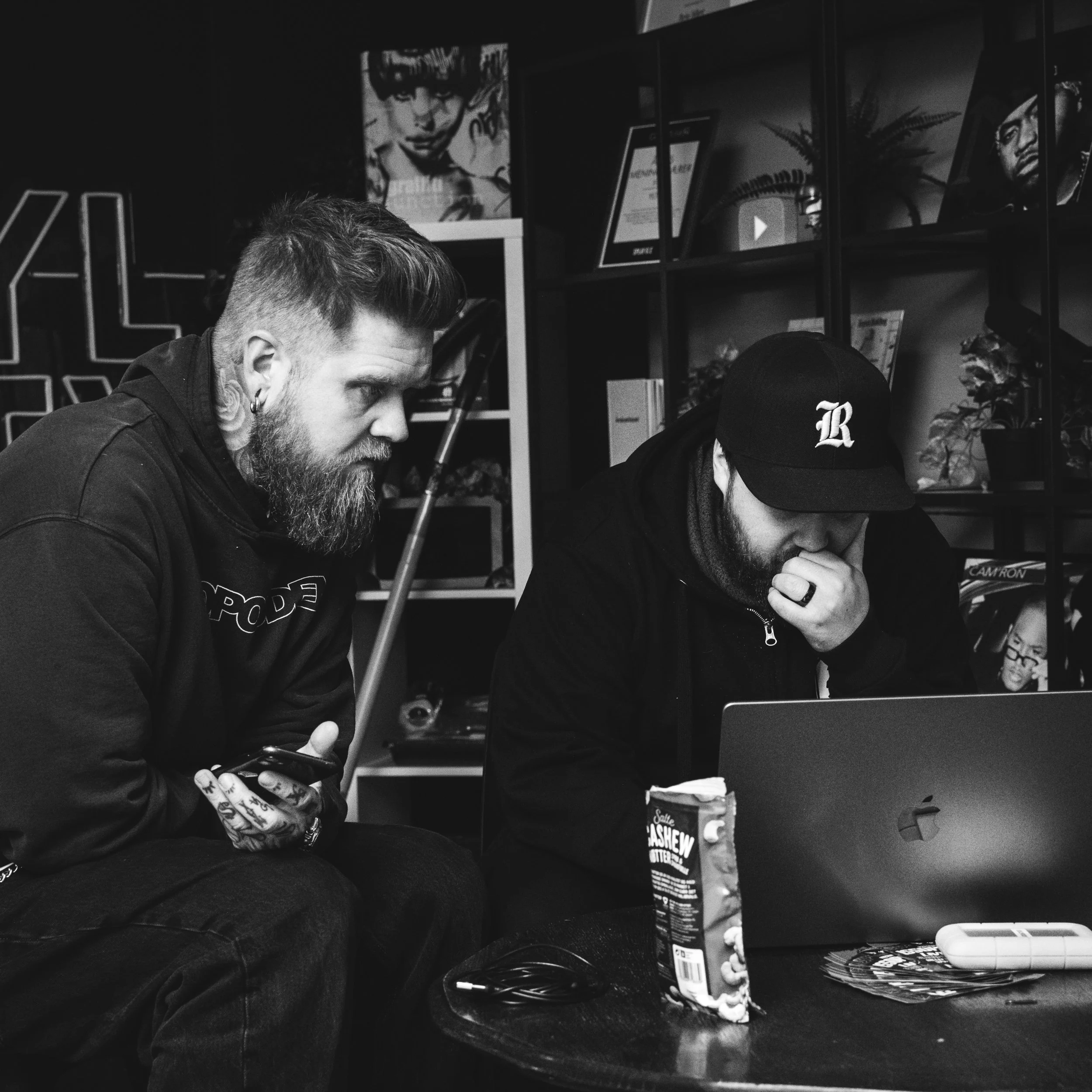 To menn ser på en laptop i en innendørs setting med hyller med bilder og planter i bakgrunnen. Den ene mannen holder en telefon og har tatoveringer på hendene, den andre holder seg til ansiktet.