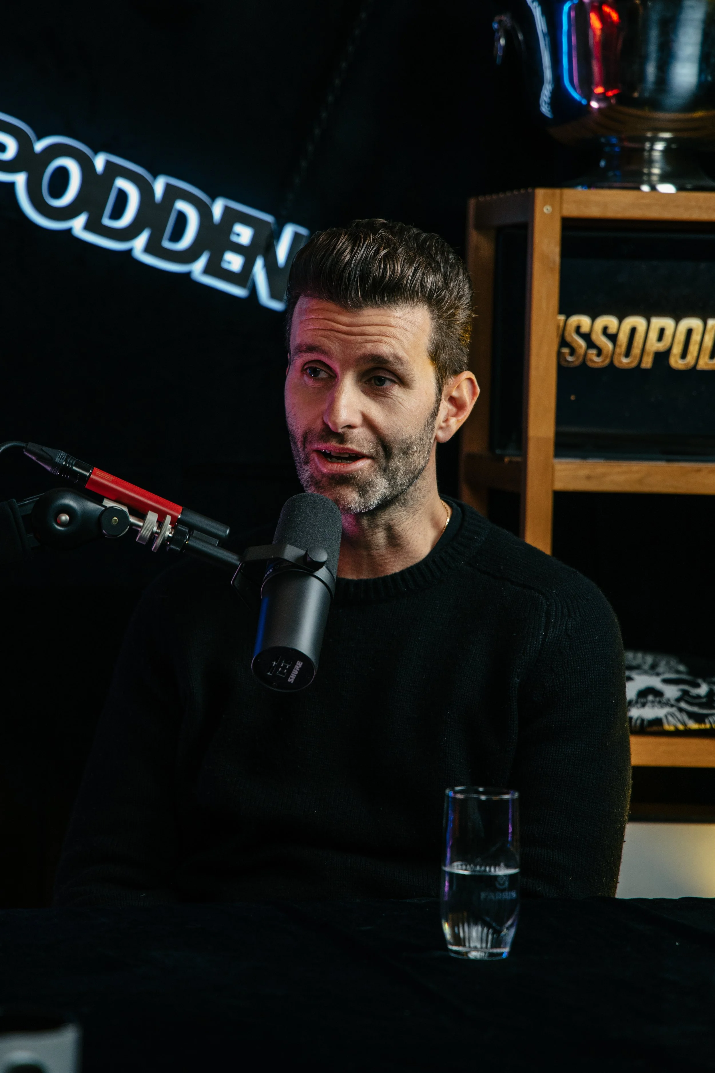 En mann med mørkt, grettent hår og skjegg snakker inn i en mikrofon under et intervju eller en podcast. Han sitter ved et bord med et glass med vann foran seg. Bak ham er en hyller med noen gjenstander og en vegg med neon-lys som har ordet 'GODDEN' o