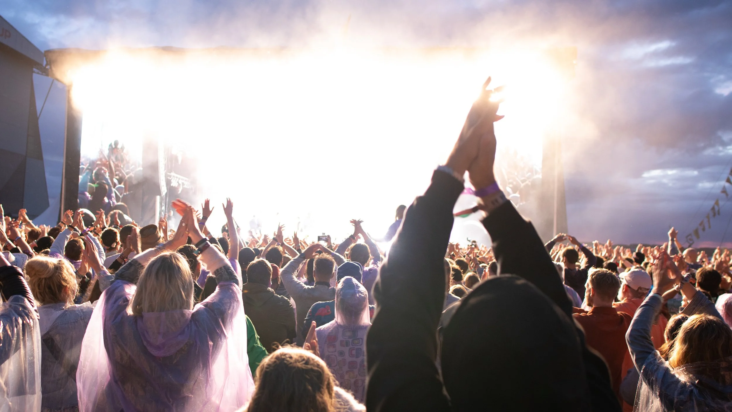 Crowd of people at a concert or festival, with their hands raised, facing a brightly lit stage with smoke and stars in the sky.