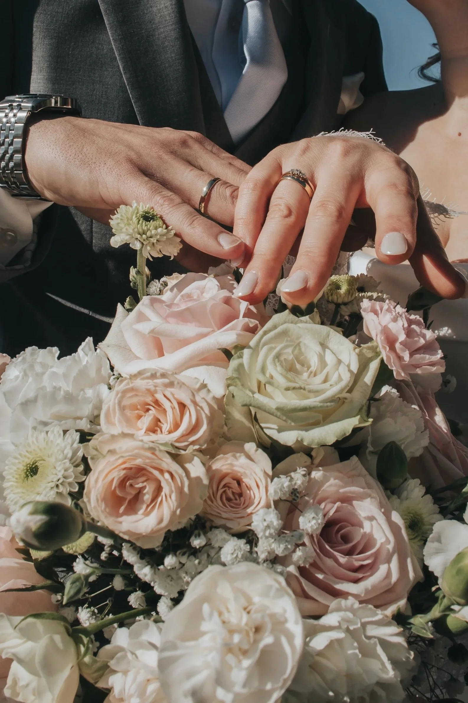 Bildet viser to personer som legger hendene sine sammen, begge bruker vielsesringer. Den ene personen har en blomsterkrans rundt hånden, og de ligger på en blomsterbukett med hvite og lyserosa blomster.