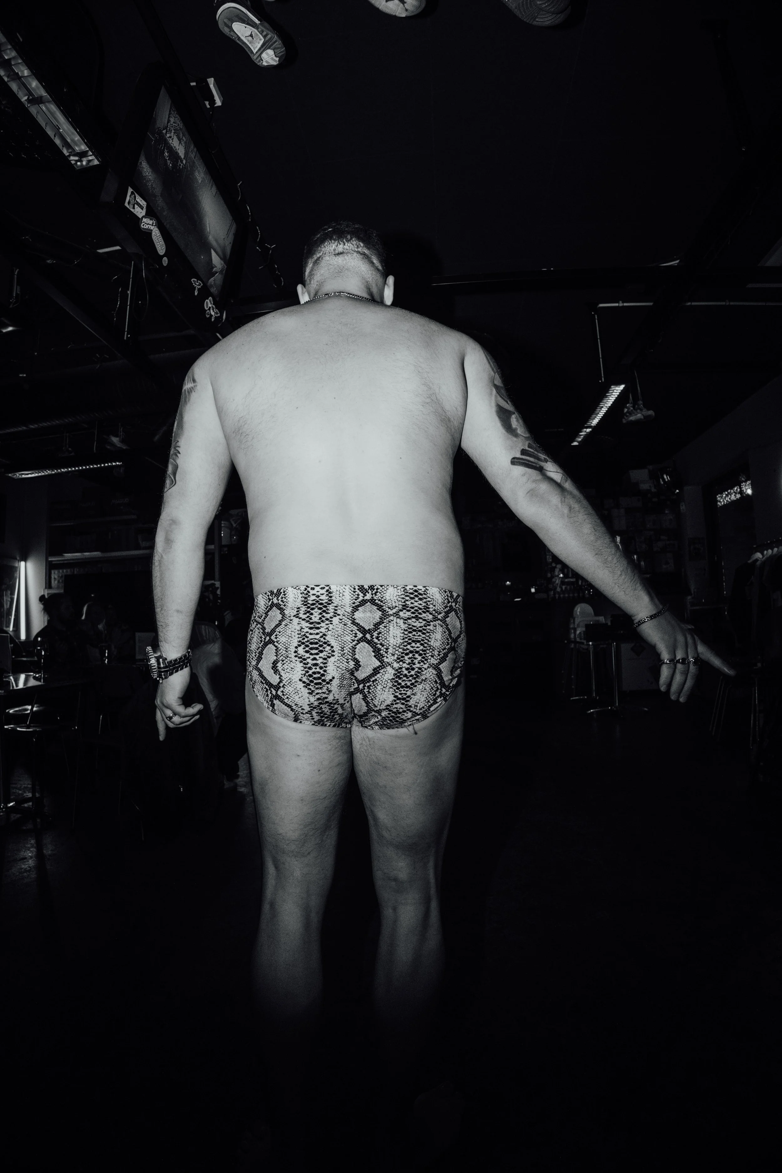 En mann med rygg mot kamera, naken fra midjen og opp, med snakeprint shorts, i en mørk bar eller nattklubbmiljø.