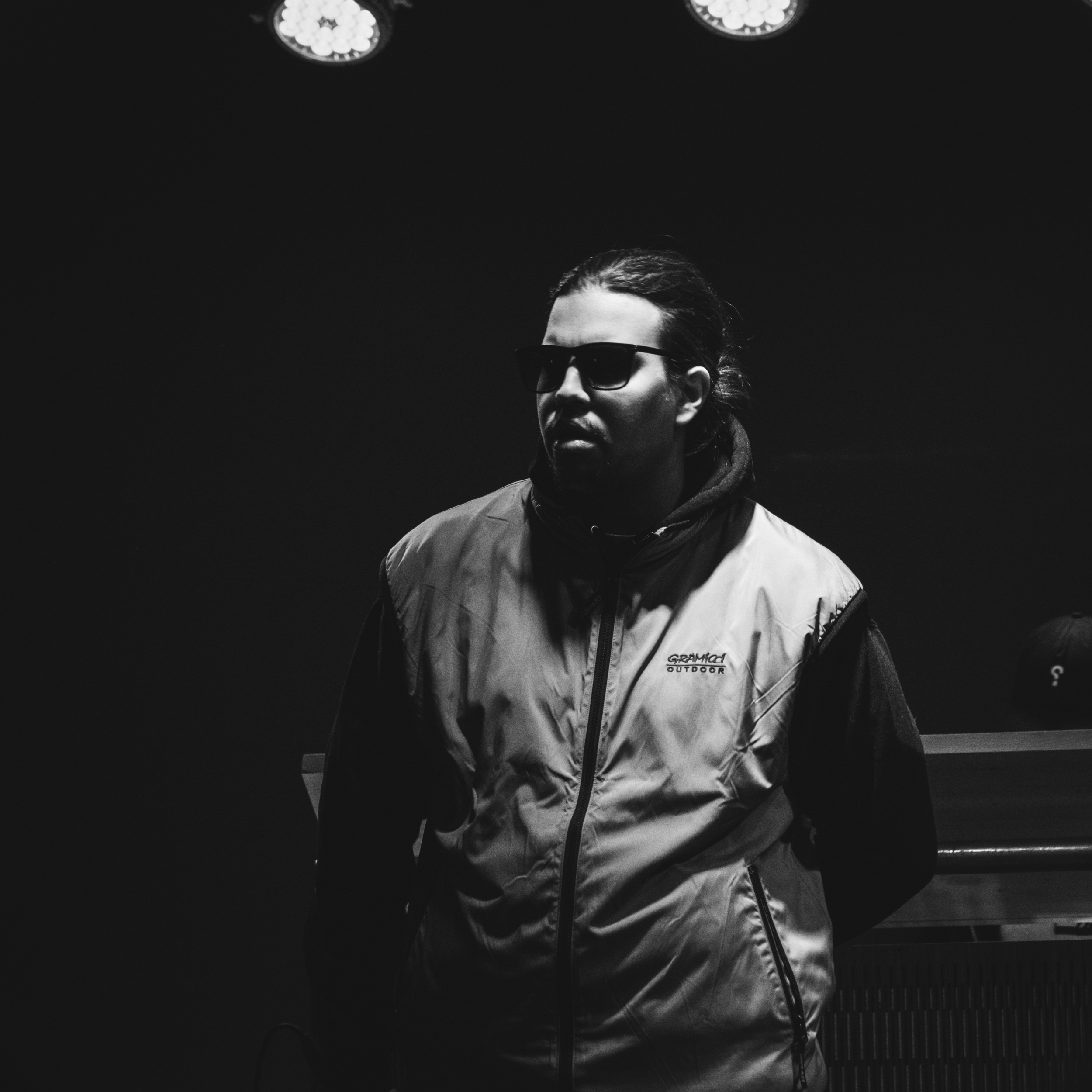 A man wearing sunglasses and a outdoor vest stands in a dark room with his hands behind his back.