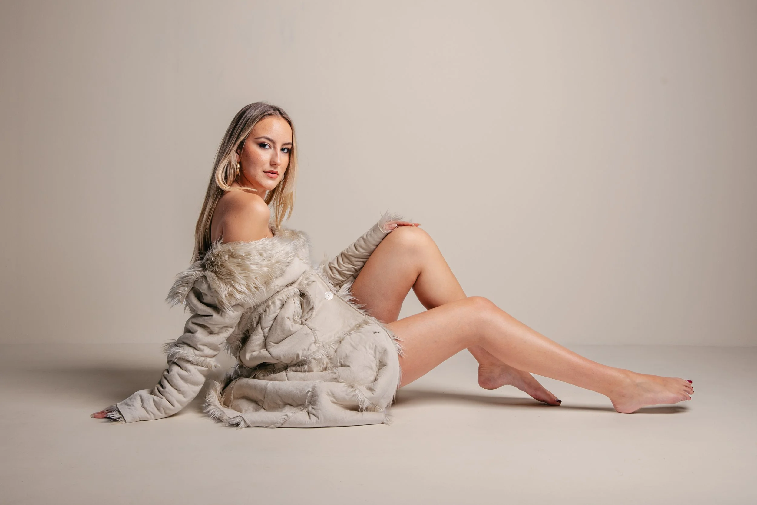 A woman with blonde hair and hoop earrings is sitting on the floor against a plain background. She is wearing a beige, fluffy coat off her shoulders and has a confident expression.