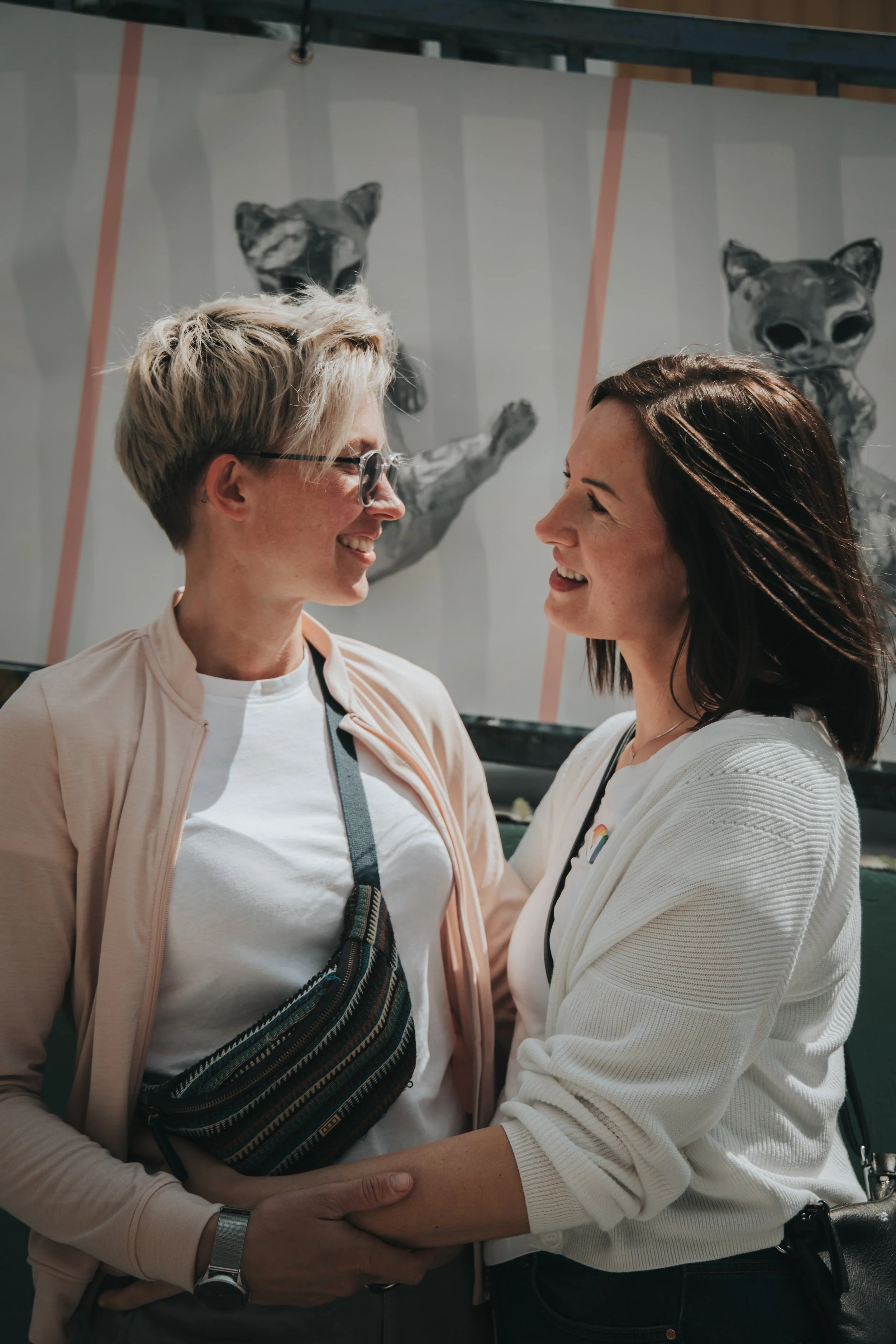 Two women are smiling and holding hands, facing each other, in front of a background with animal posters.