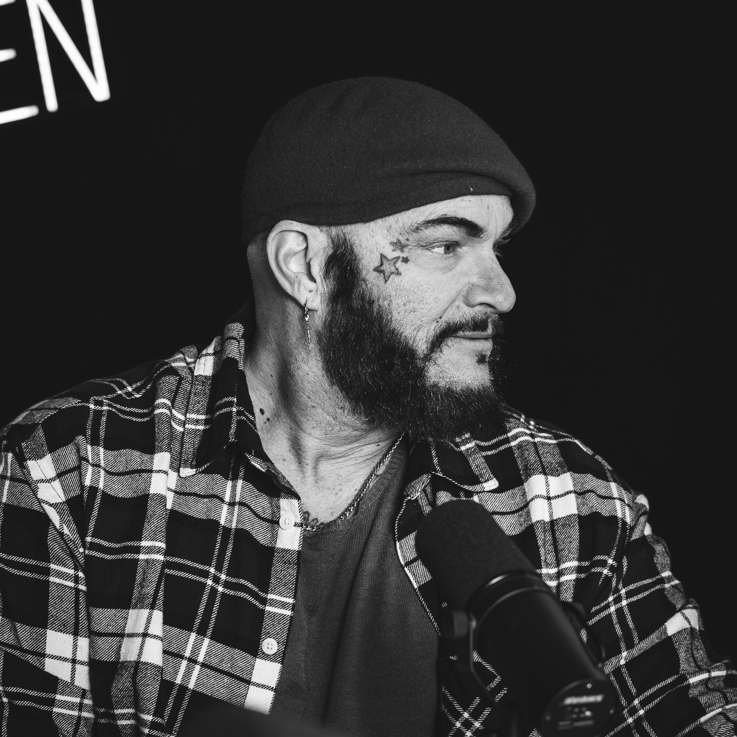 En mann med skjegg, earring og tatovering av en stjerne under øyet, iført en svart lue og sjall med rutemønster, snakker inn i en mikrofon.