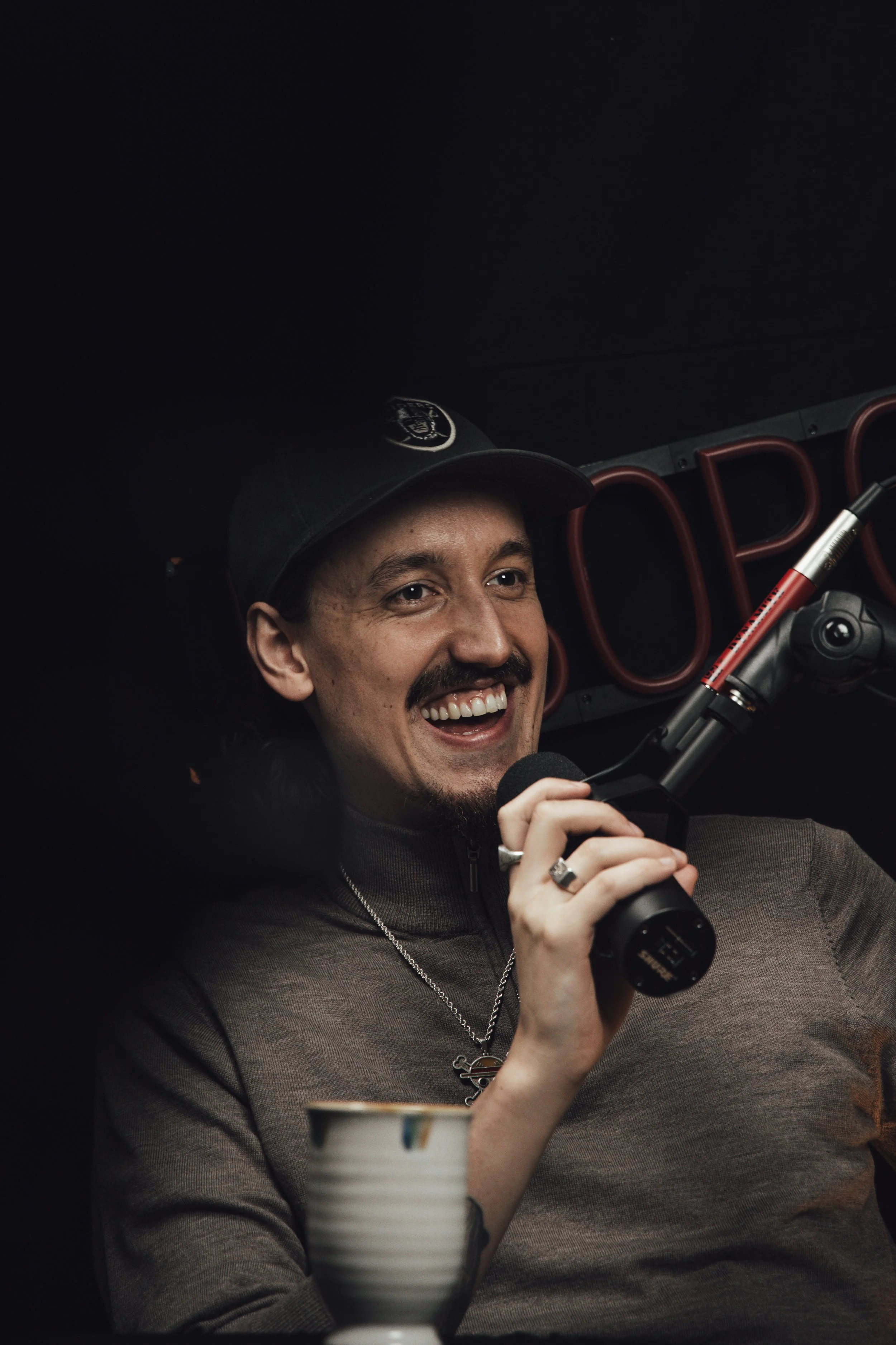 Mann med skjegg og hat smilende, snakker inn i mikrofon, student eller vlogger, mørk bakgrunn med rødt neonskilt.