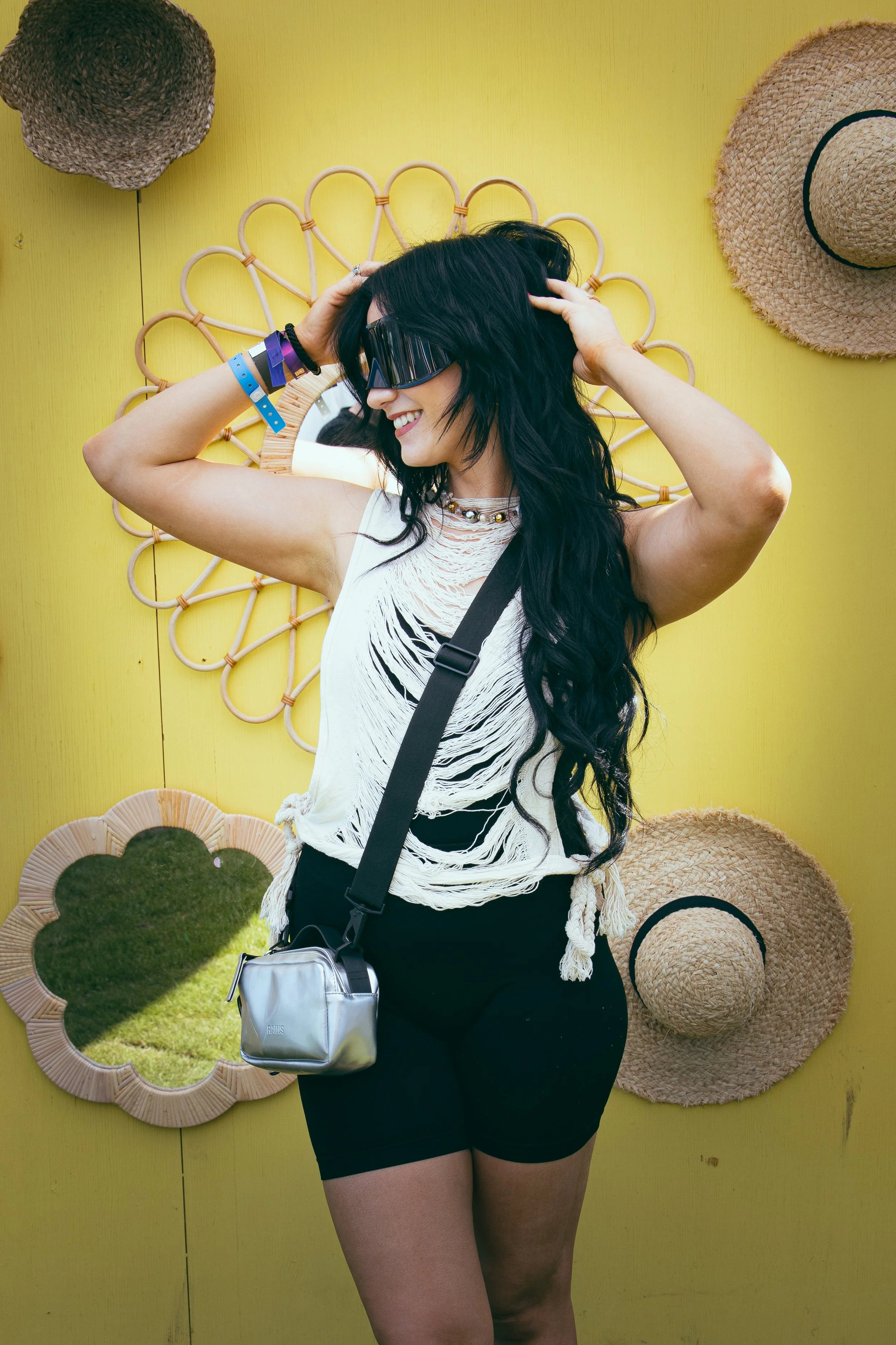 En kvinne med langt, mørkt hår, smilende, iført solbriller og sommerklær, står foran en gul vegg med speil og hatter.