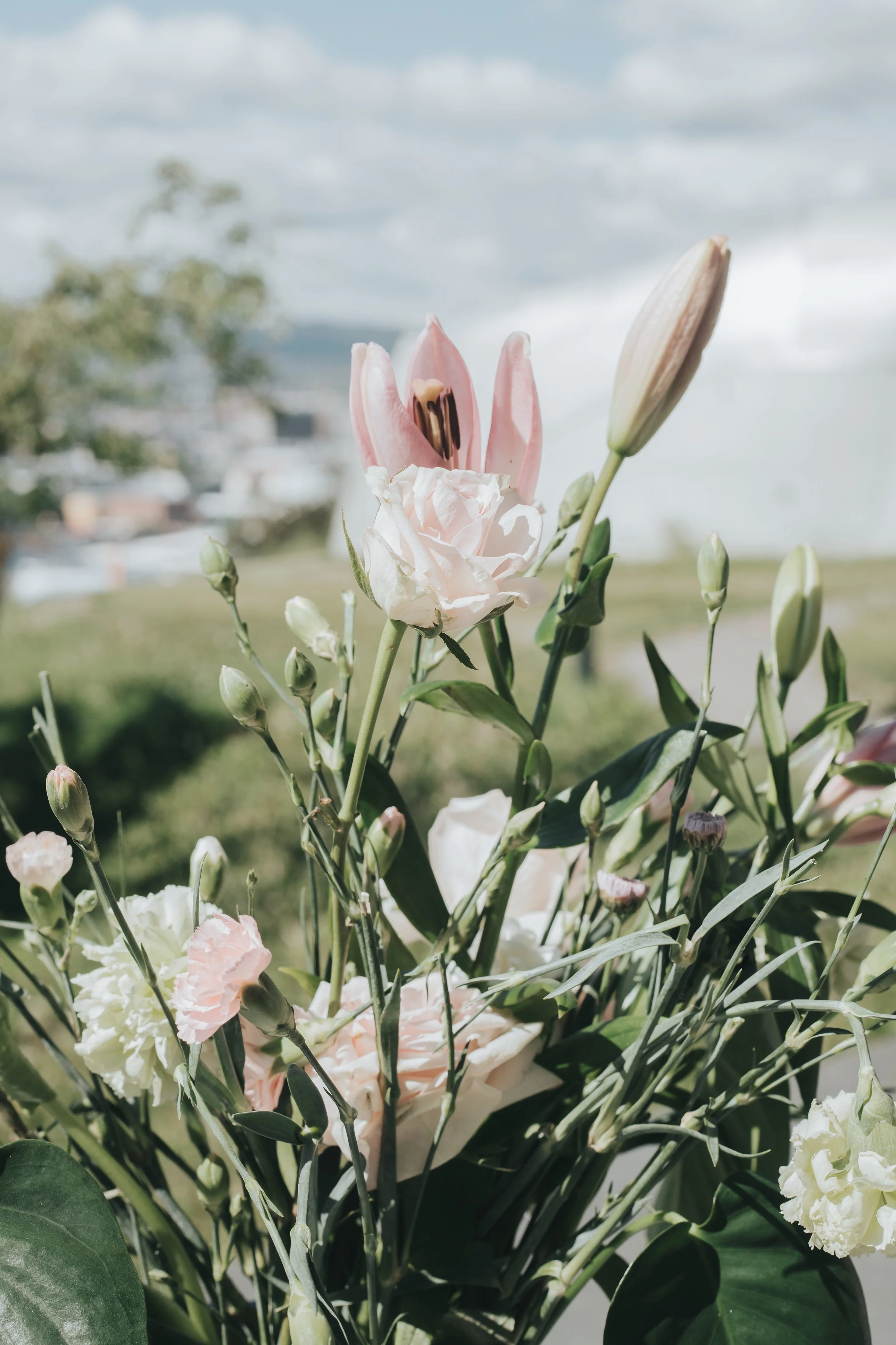 En bukett av rosa og hvite blomster, inkludert en lilje, mot en utendørs bakgrunn med blå himmel og skyer.