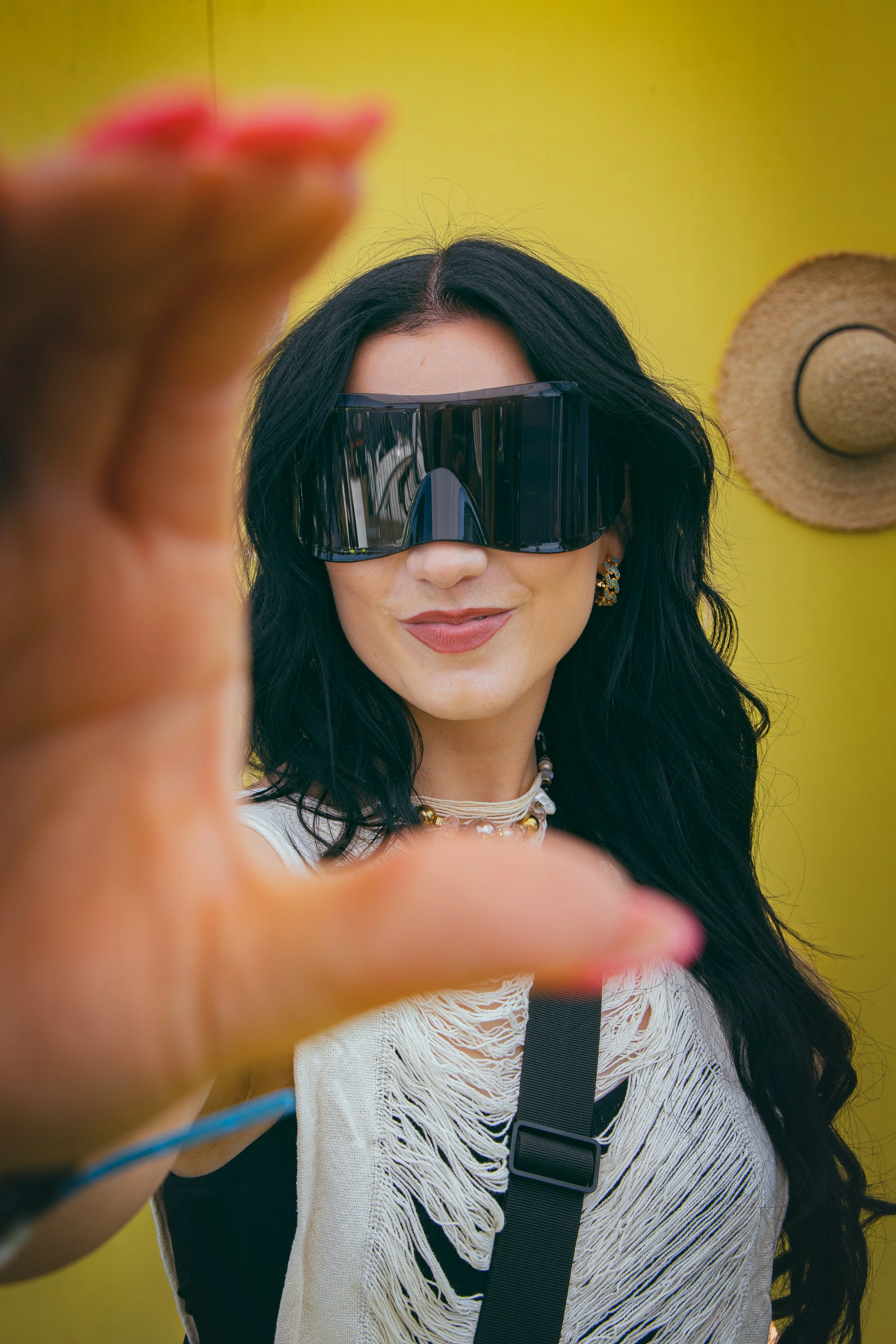 Woman with long black hair wearing large black sunglasses, gold earrings, layered necklaces, and a distressed white shirt, standing against a yellow background with a tan hat hanging on the wall.