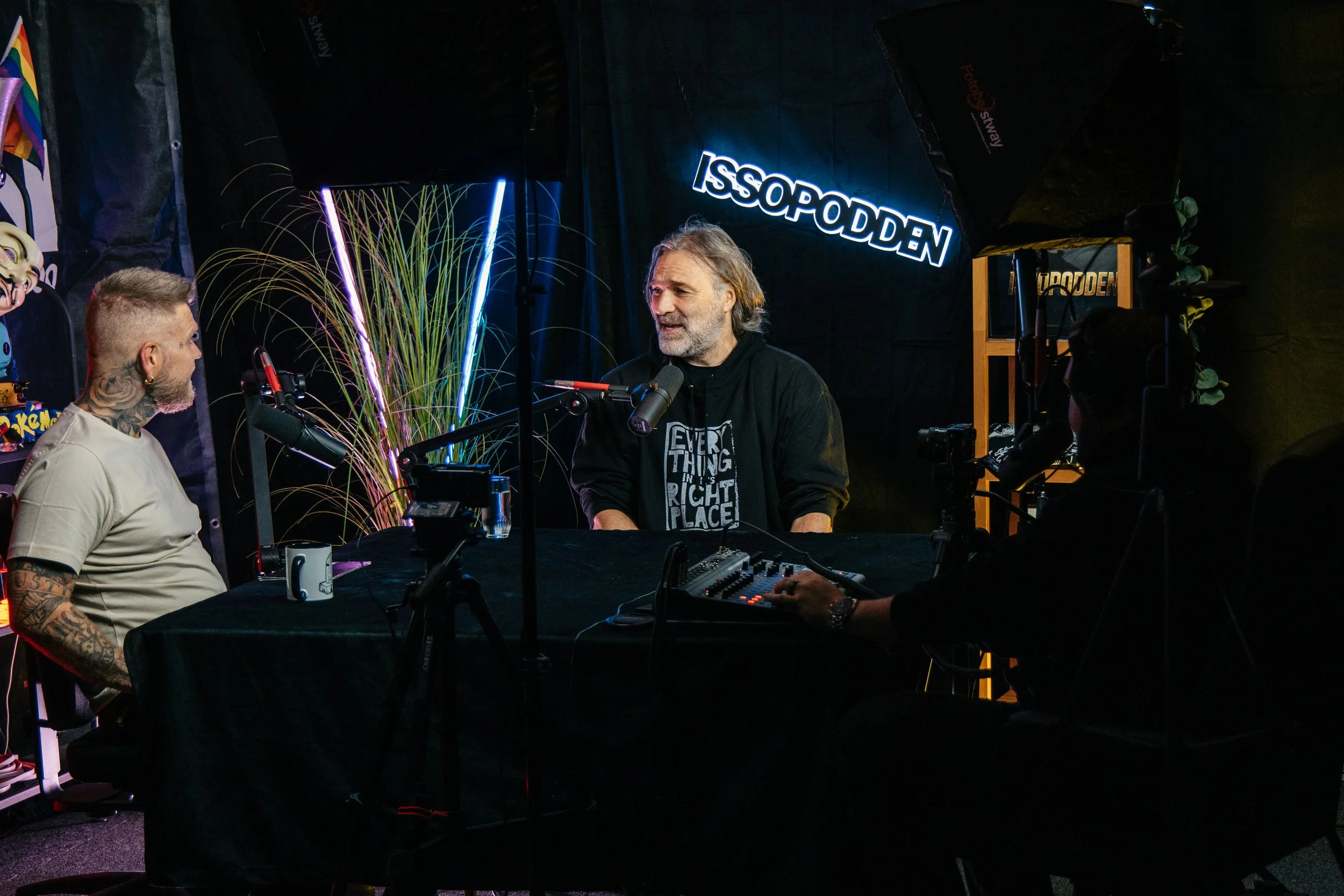 To personer deltar i en podkastsamtale på et mørkt studio med en bakgrunn som har navnet 'ISSO GODDEN' opplyst i neon. Den ene personen har lys hud, grått hår og skjegg, og snakker inn i en mikrofon. Den andre er en mann med tatoveringer, kort hår, o