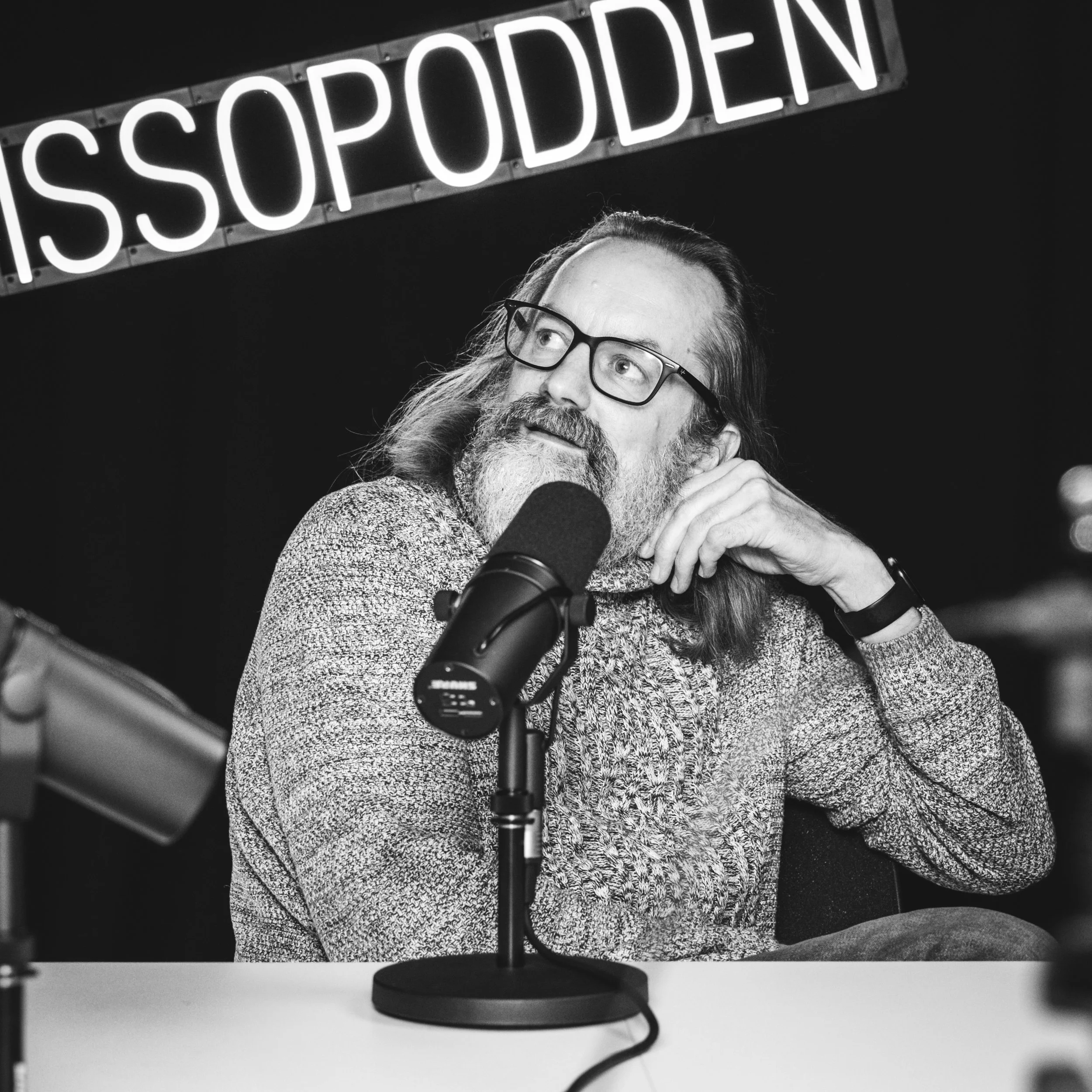 En mann med briller og lang hår, som har skjegg, sitter foran en mikrofon i en plate-studio. Bak ham er det en skilt med teksten "ISSOPODDEN". Bildet er i svart-hvitt. Egil Bare Egil