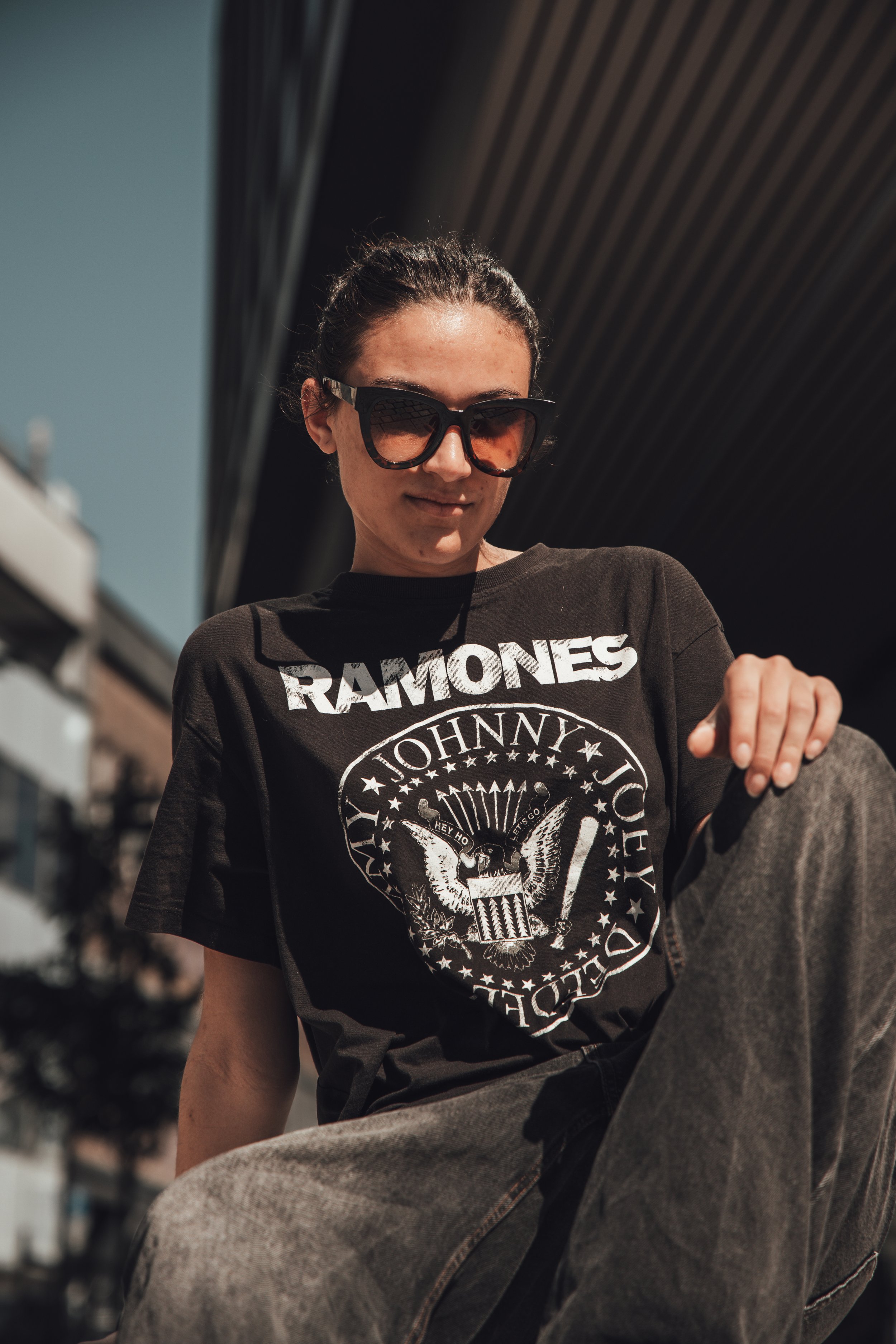 En kvinne iført solbriller og en svart Rock band T-skjorte med tekst og grafikk, utendørs under solen.