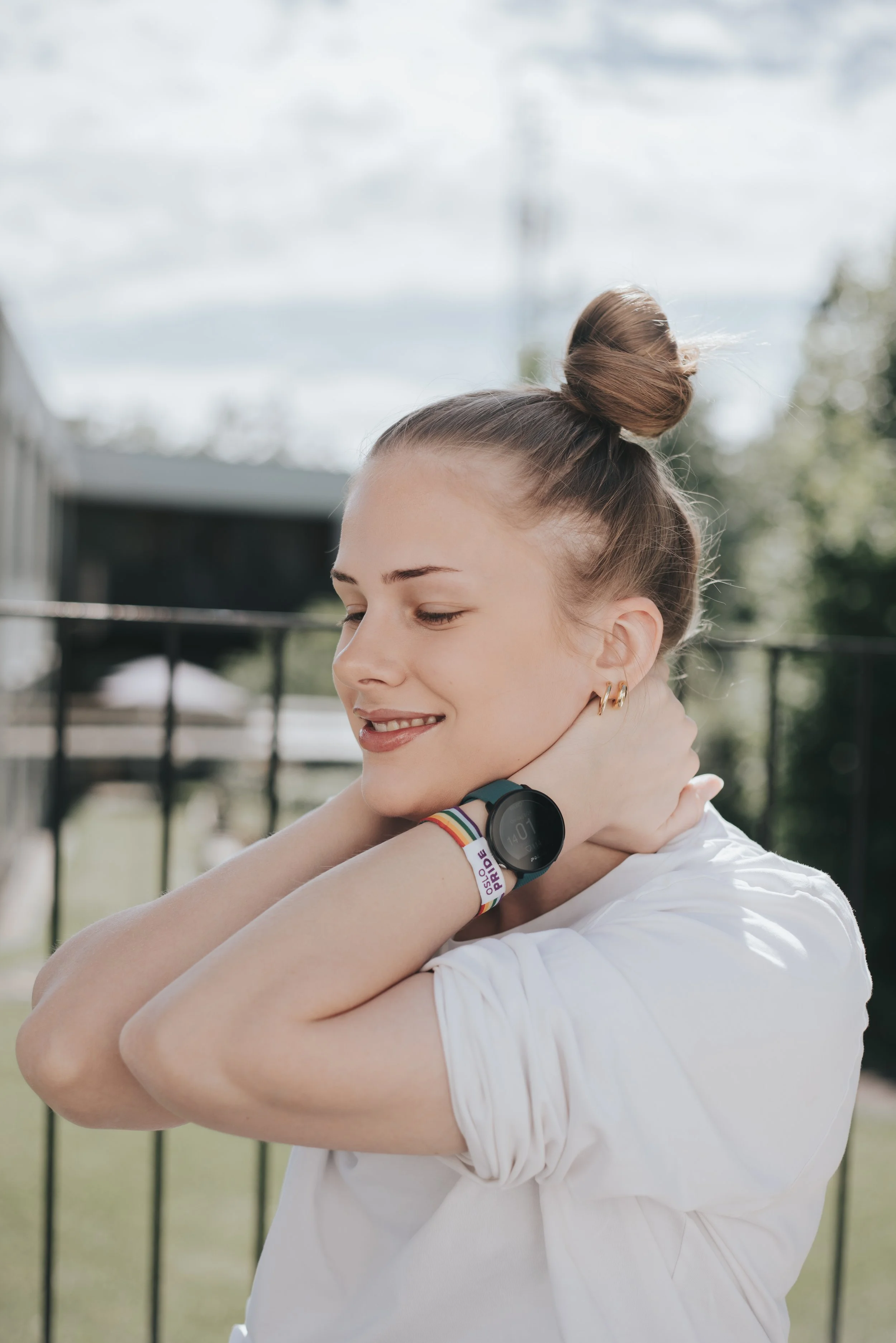 Ung kvinne med håret i en høy knute, smiler med øynene lukket, i en utendørs setting, bærer en svart klokke og en fargerik armbånd. Her har hun armene rundt nakken til en annen person, bakgrunn med grønt landskap og en svart gjerde.