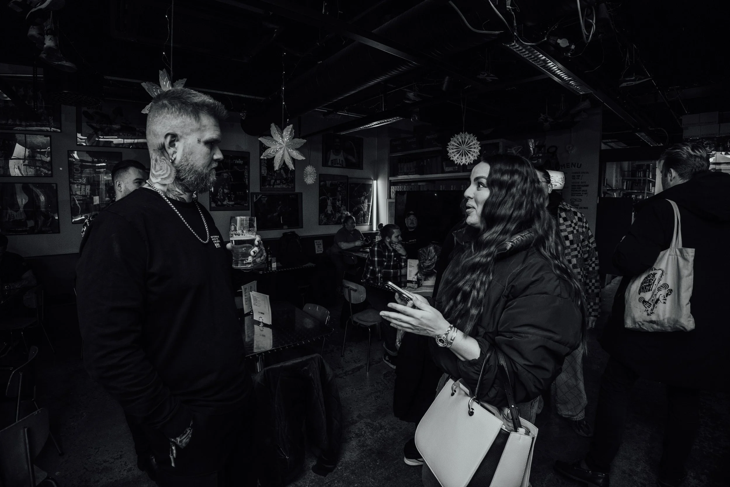 To personer snakker i en bar. Den manlige personen har tatoveringer, krøllete hår og holder et glass med drikke. Den kvinnelige personen har lang, bølget hår og bruker en svart jakke, og hun holder en telefon.