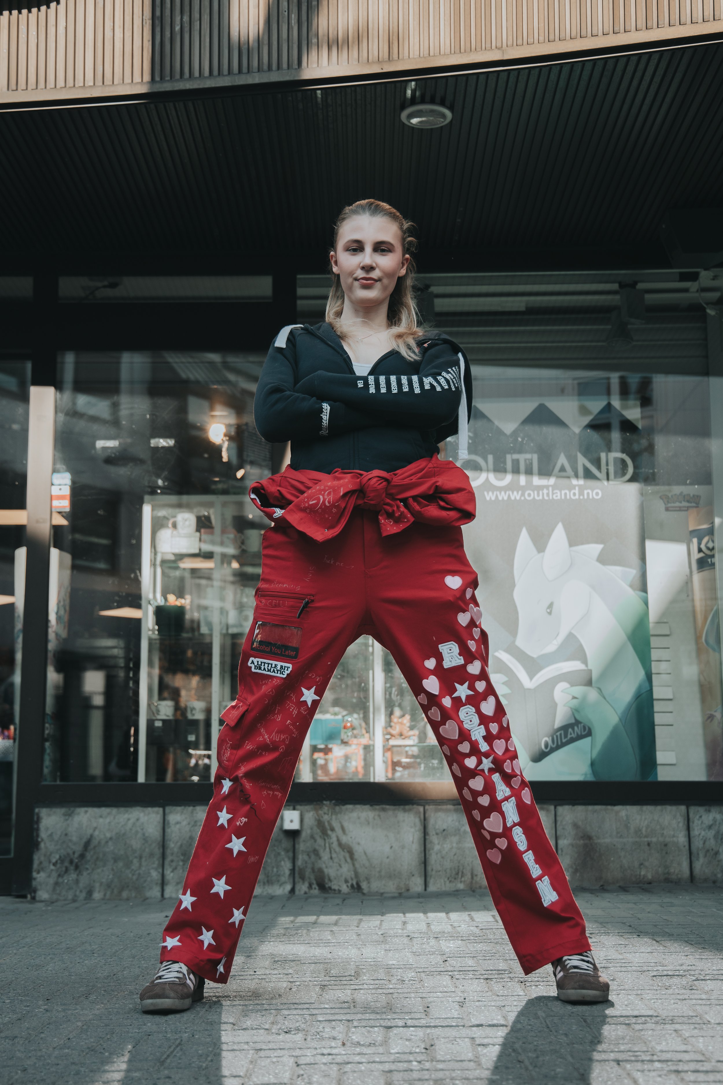 En kvinne står med bena i skulderbredde, krysser armene og ser direkte mot kamera foran en butikkfront med glassvinduer og utendørs dekorasjon.