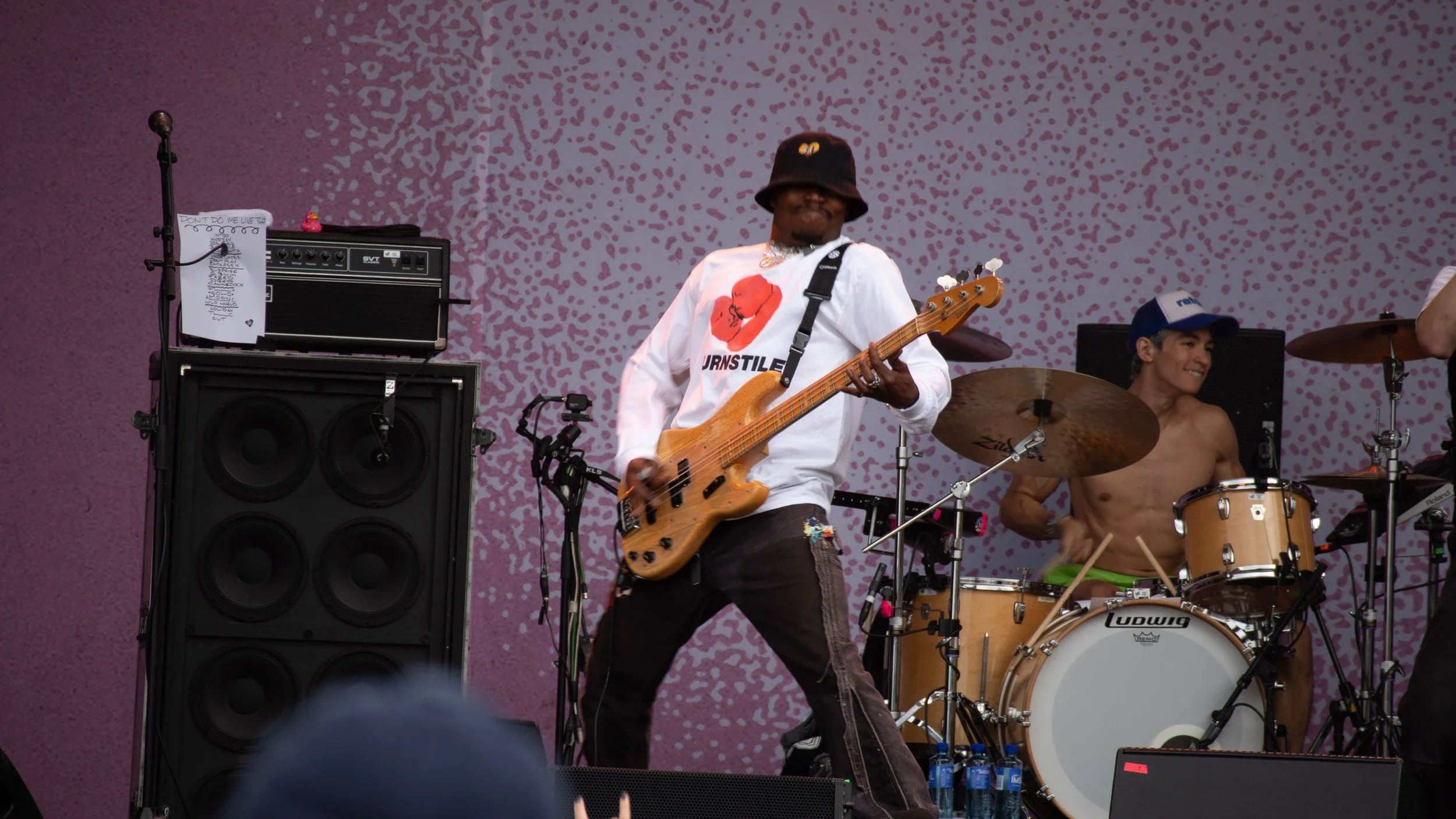 Musiker spiller bassgitar og trommeslager uten skjorte under en konsert på en scene med rosa og lilla bakgrunn.