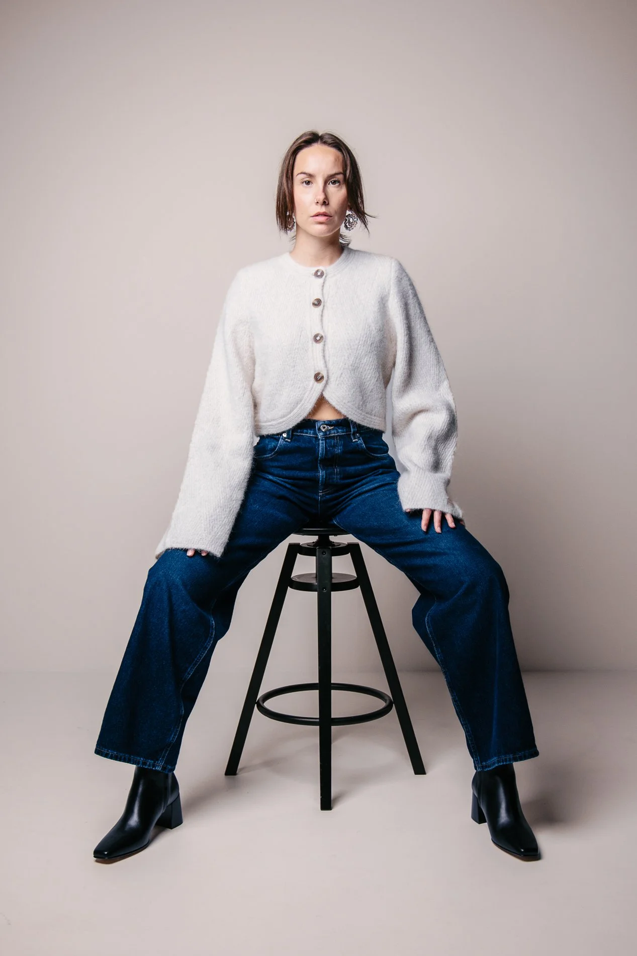 A woman sitting on a stool against a plain background, wearing a cream-colored cardigan, dark blue jeans, and black heeled boots.