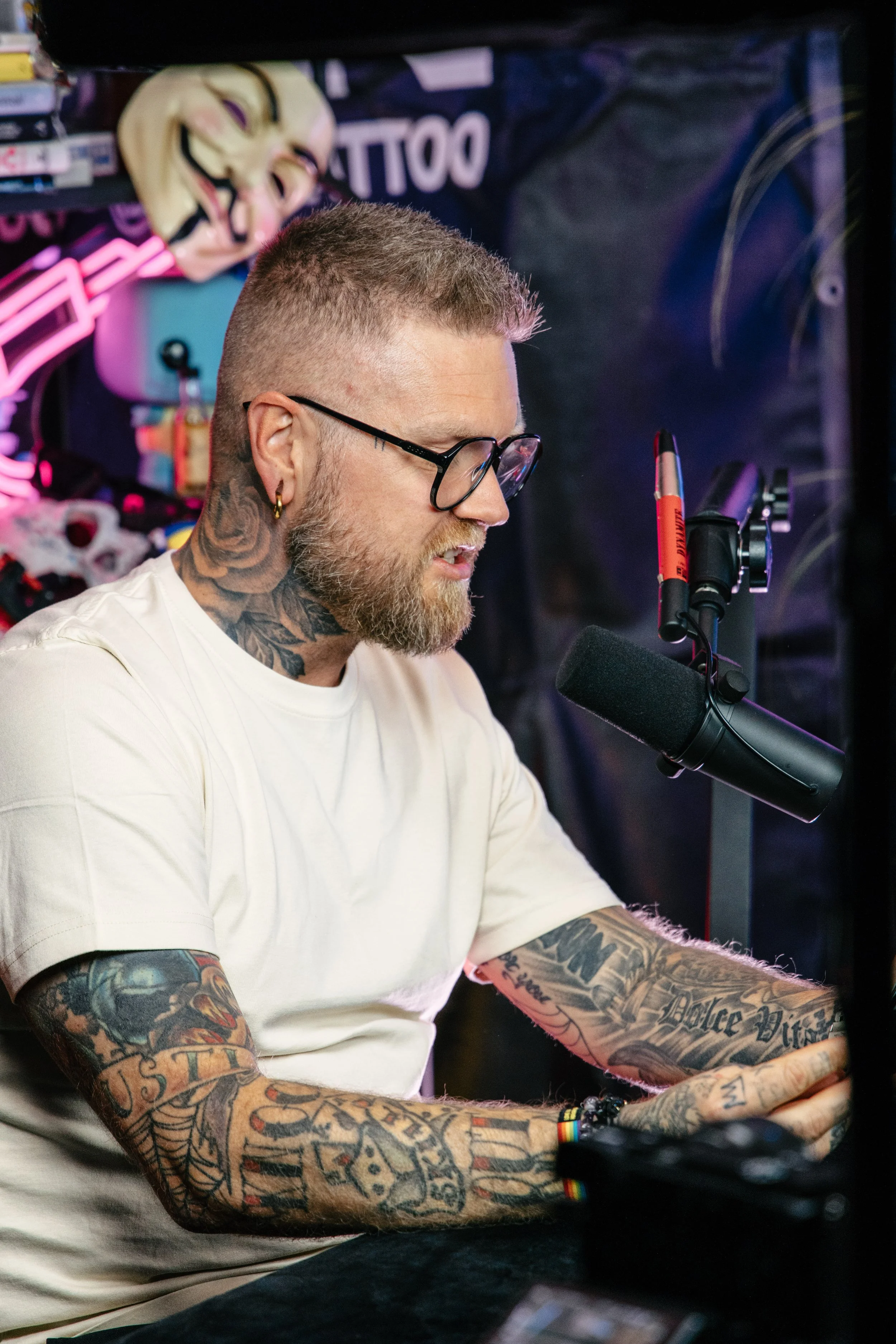 En mann med tatoveringer, briller og skjegg som snakker inn i en mikrofon i et studioumråde med neonbelysning og et Guy Fawkes-masker.