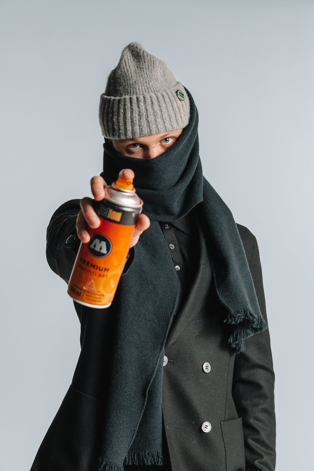 A person wearing a gray knit beanie, a black scarf covering their lower face, and a black coat, holding a can of spray paint towards the camera.