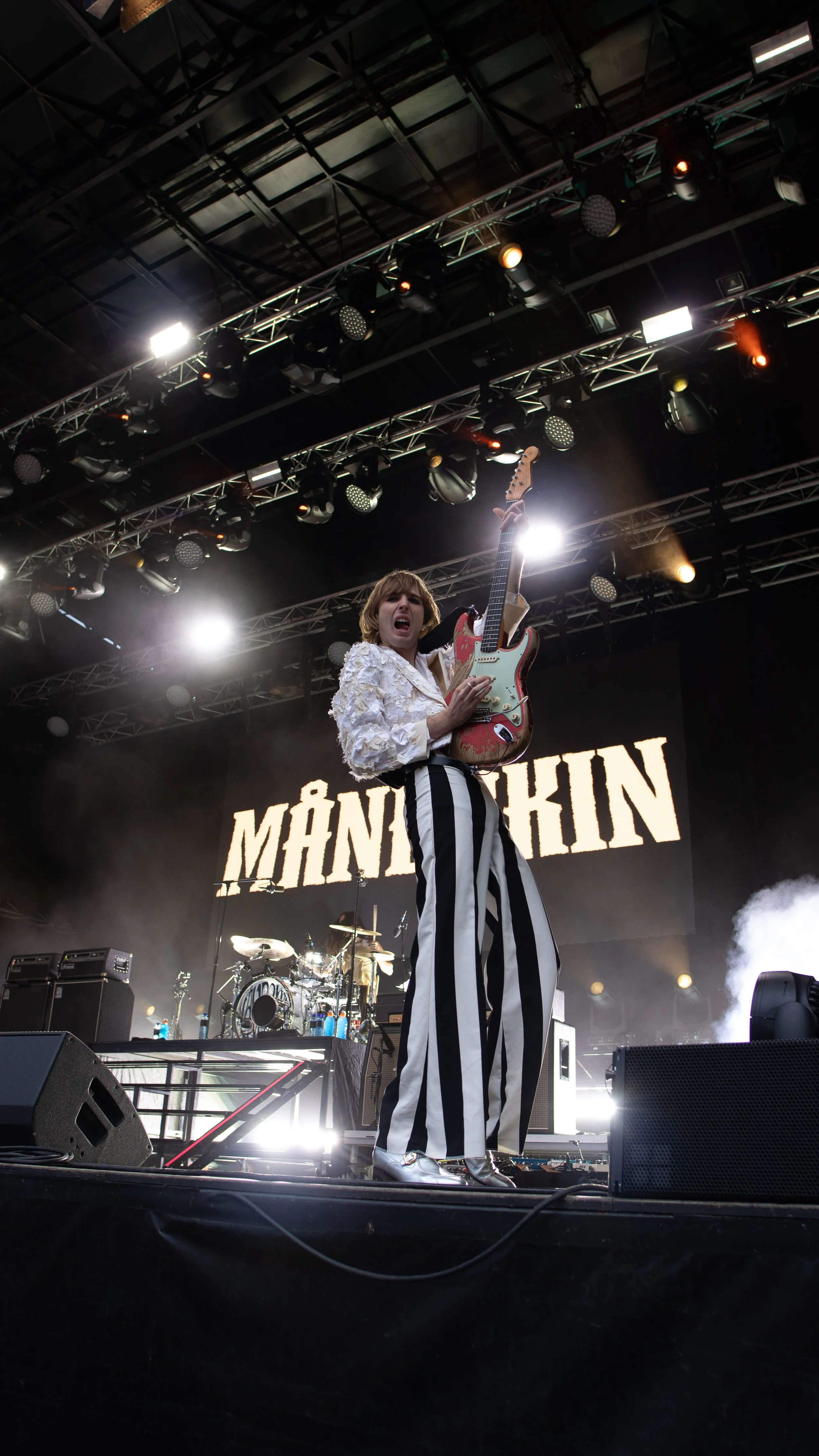 En kvinne på scenen under en konsert, holder en elektrisk gitar opp i luften, med navnet MÅNICHIN i bakgrunn. Scenelys og lydutstyr er synlig.