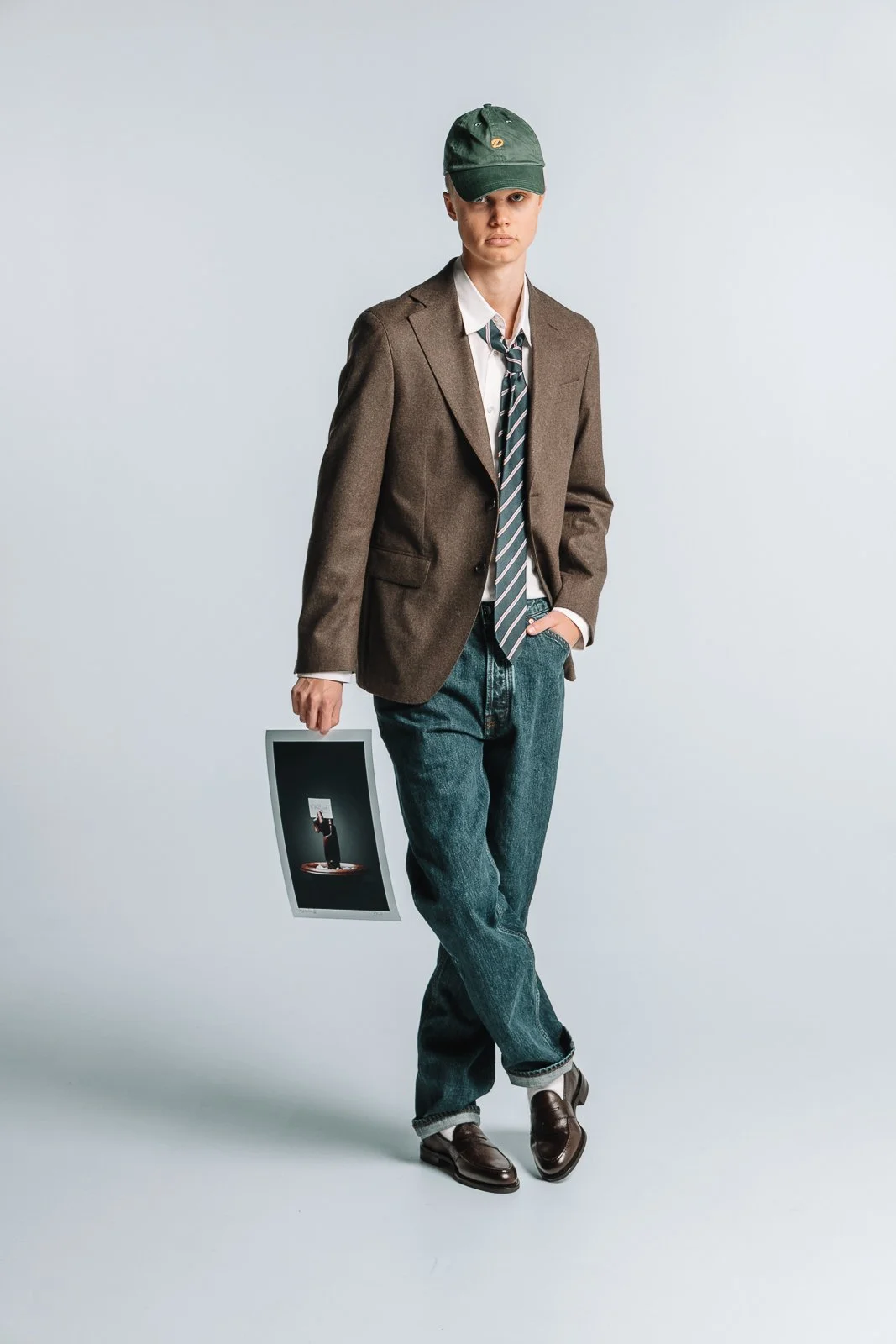 Person iført en brun blazer, hvit skjorte, stripet slips, blå jeans og brune sko, holder et fotografi i hånden.