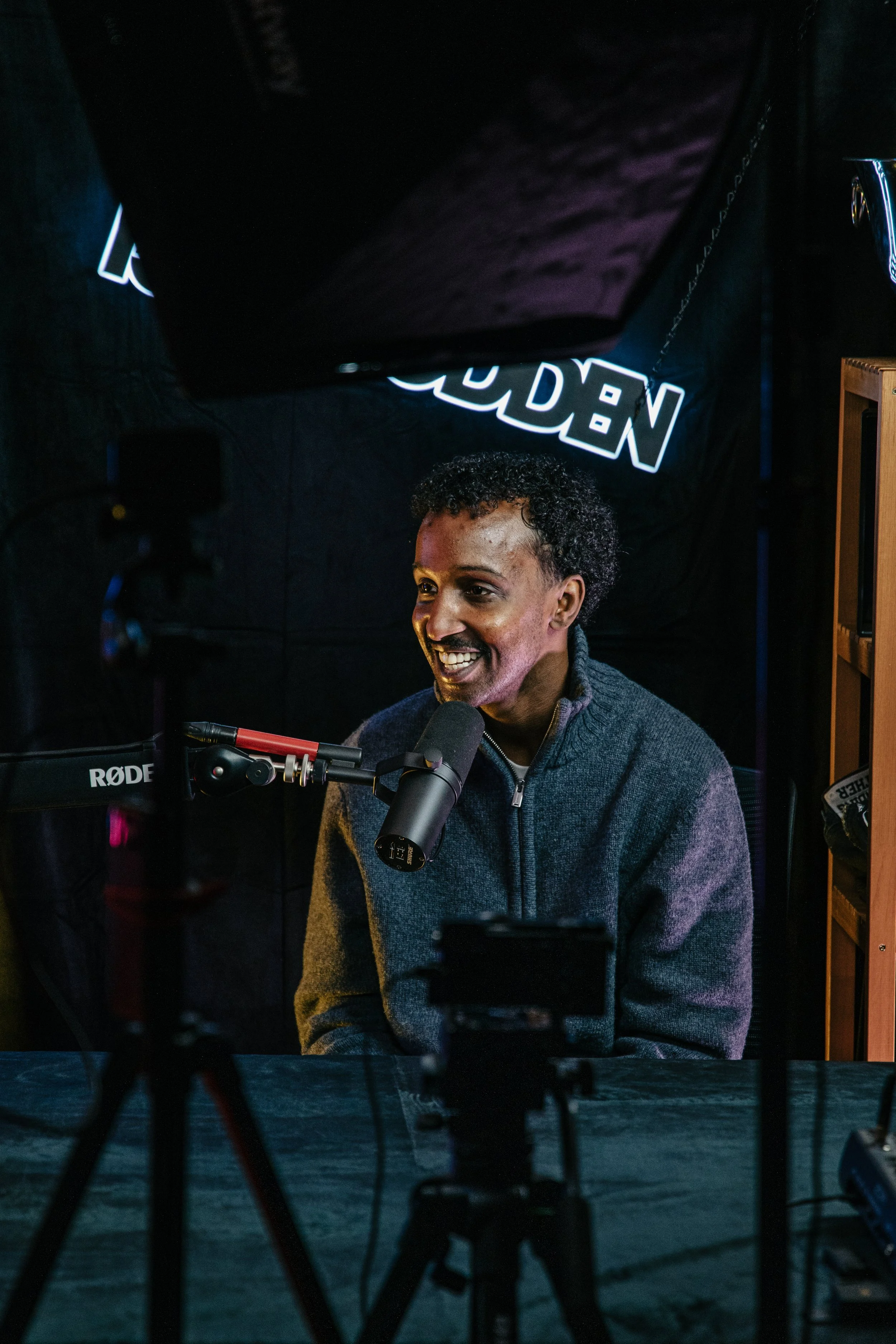 En mann med krøllete svart hår, smilende, sitter foran en mikrofon i et mørkt rom, med radio- eller studioutstyr rundt seg.