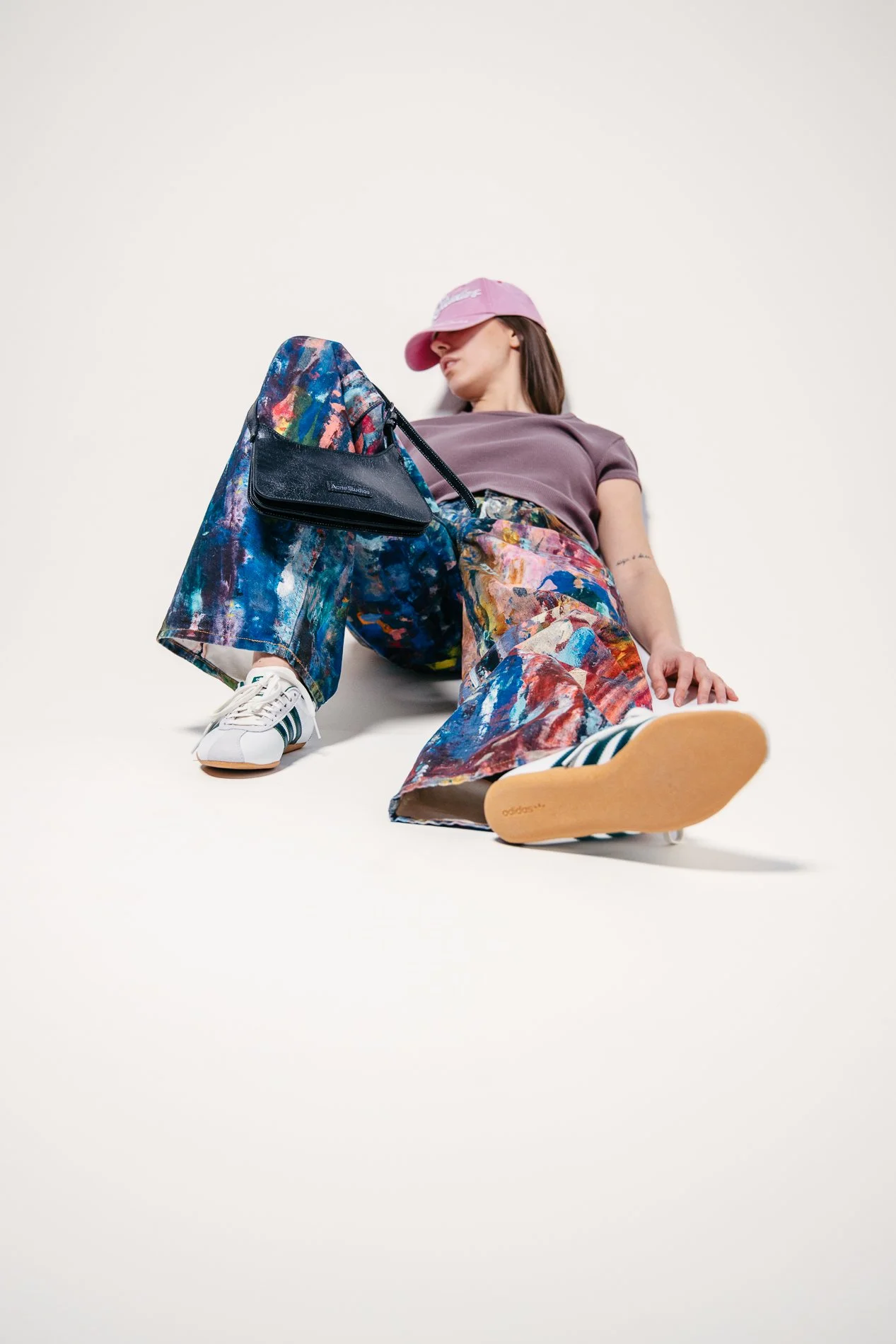 Et lavt vinkelbilde av en kvinne i fargerike bukser og en lilla t-skjorte, som sitter på gulvet med en vestlig hvit bakgrunn, med et rosa cap og Adidas sneakers med blå striper.