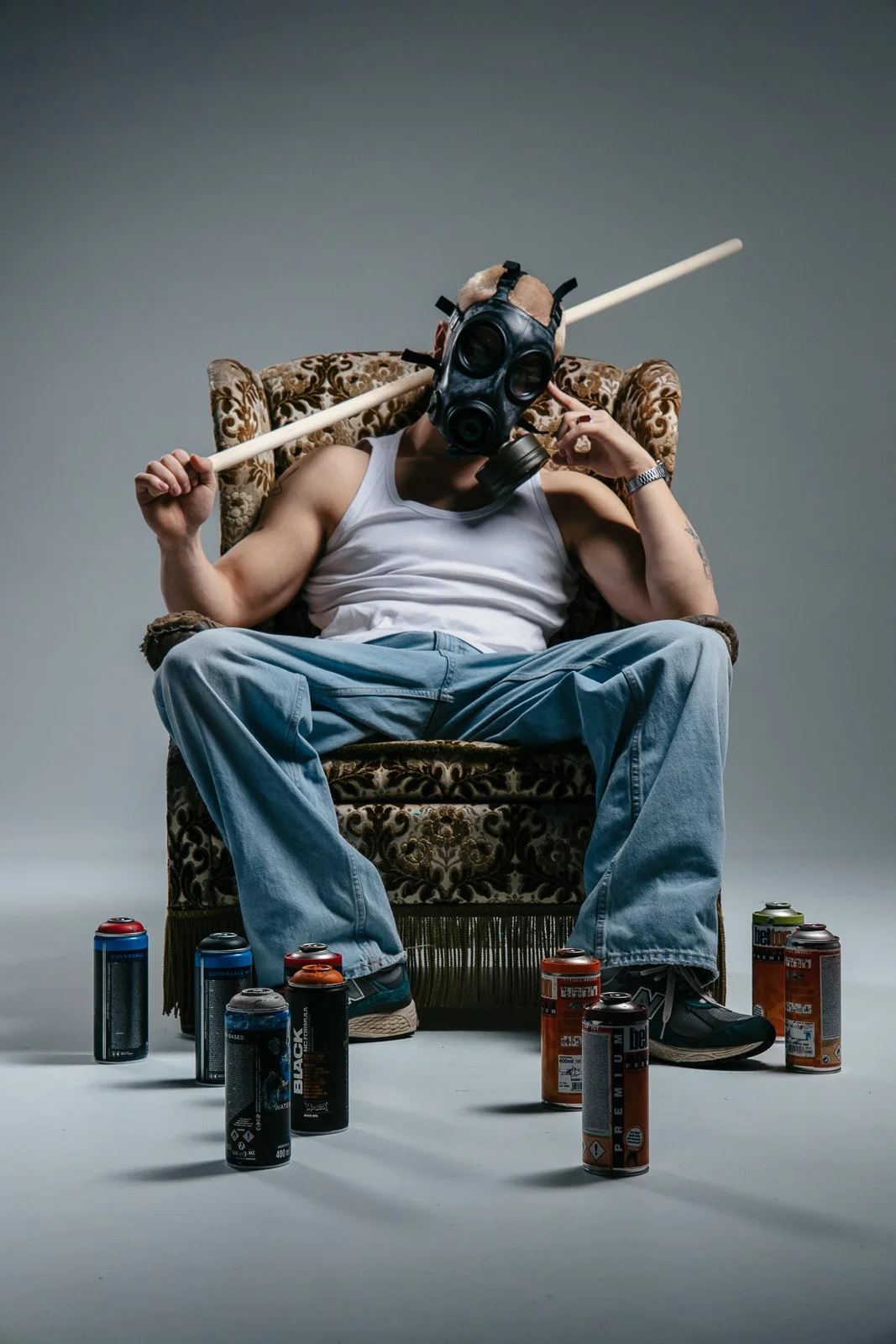 A person sitting on an ornate armchair, wearing a black gas mask and a white tank top, holding a long stick over their shoulder, surrounded by spray paint cans on the floor.