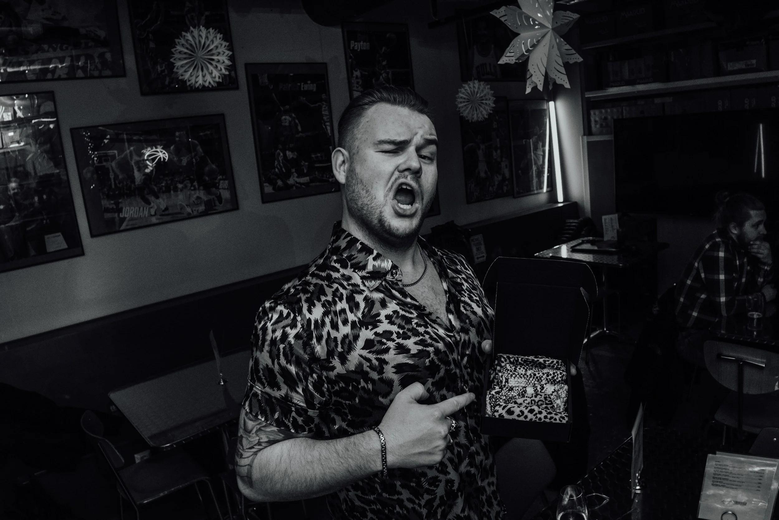 Ung mann i leopardmønstrete skjorte som gjør en dramatisk grimase og peker på seg selv, i en bar eller restaurant, svart-hvitt. Crazy ass