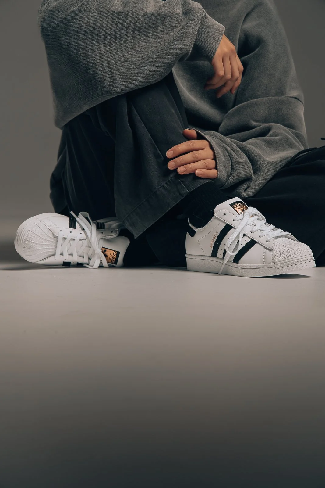 Person iført svarte sneakers med hvite og svarte detaljer, sittende på gulvet og kledd i grå genser og svarte bukser.