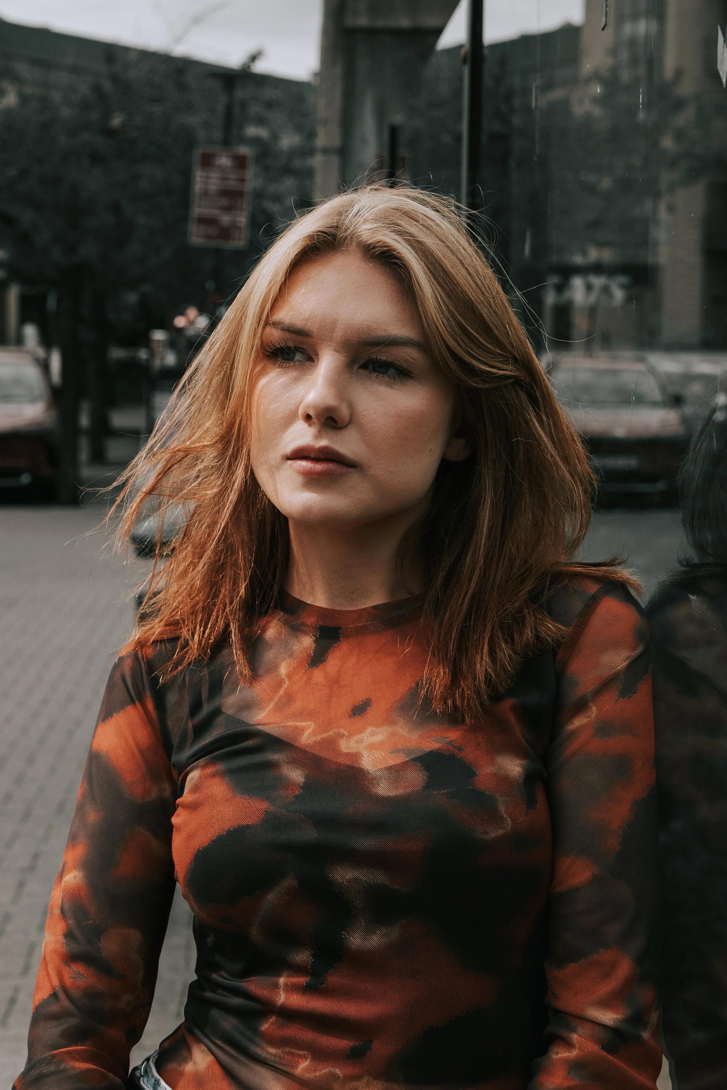 Portrett av en kvinne med brunt hår, som står ved en glassvegg på en gate, iført en mønstret rød og svart topp.