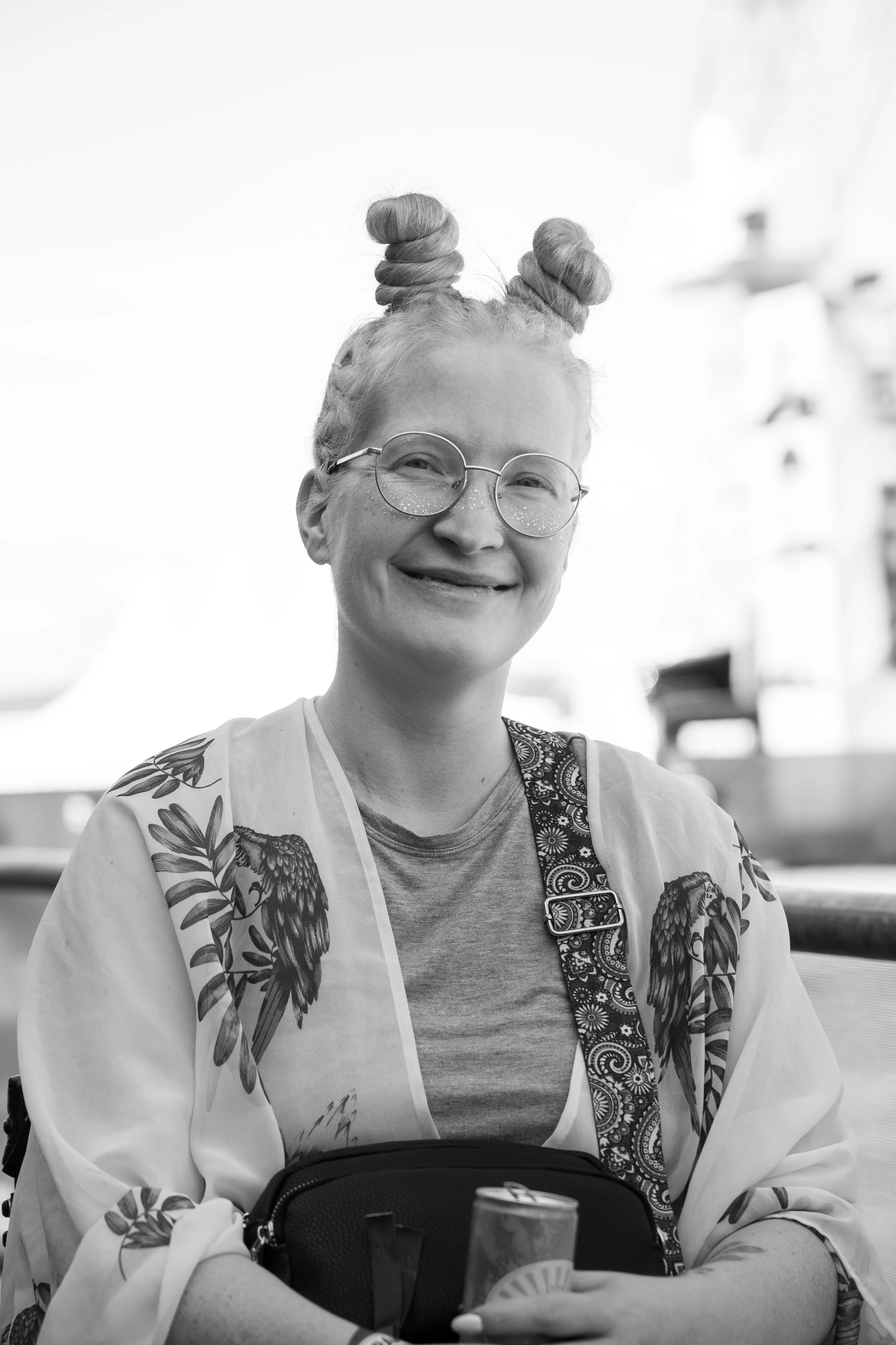En kvinne med to spiralformede hårknuter, iført runde briller med glitter, smilende, iført en t-skjorte og en kjole med fuglemønster, holder en kopp og en energidrikk, utendørs i solskinn. Slottsfjell festivalen 2025