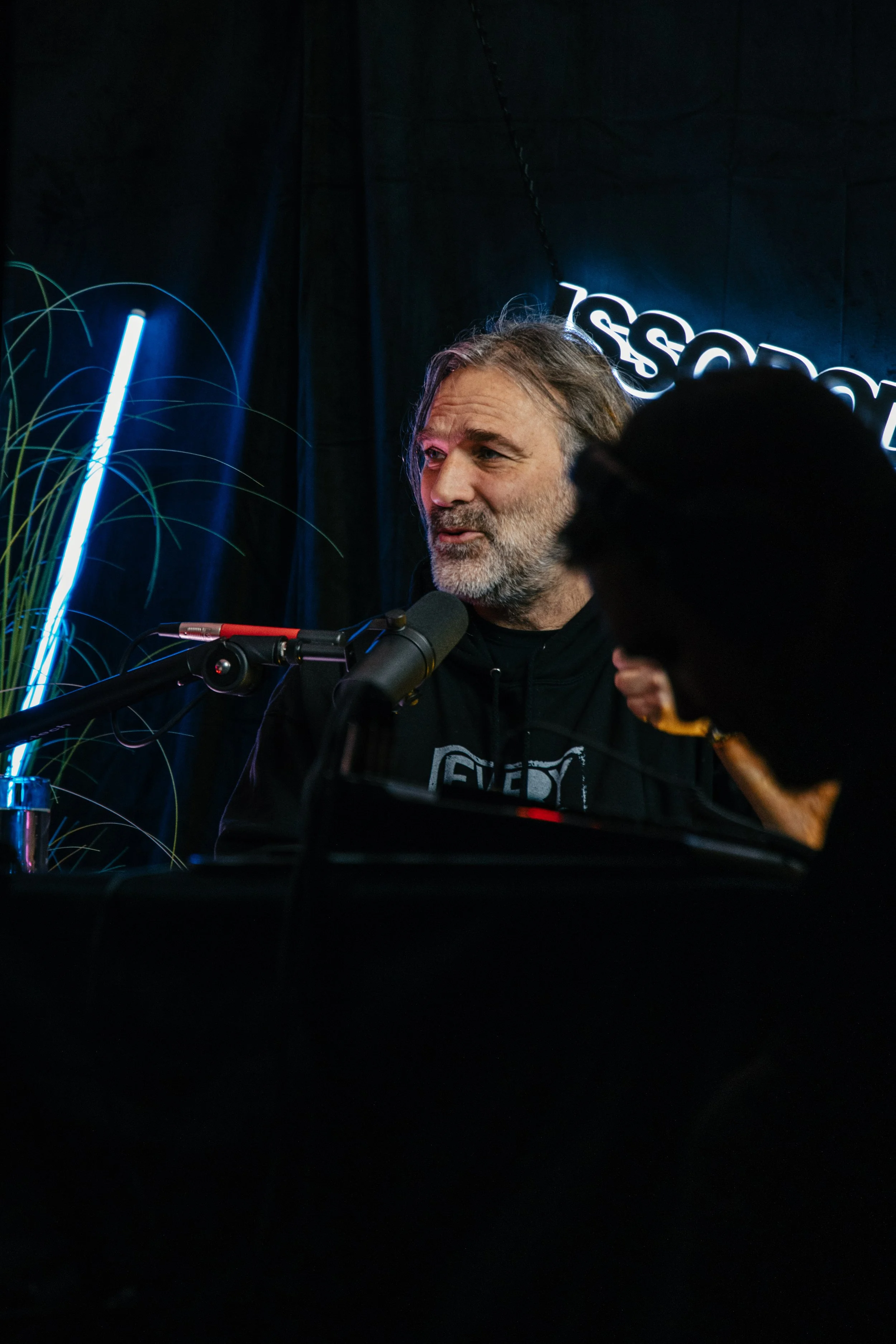 En mann med grått skjegg snakker inn i en mikrofon, omgitt av mørk bakgrunn og blå neonlys.