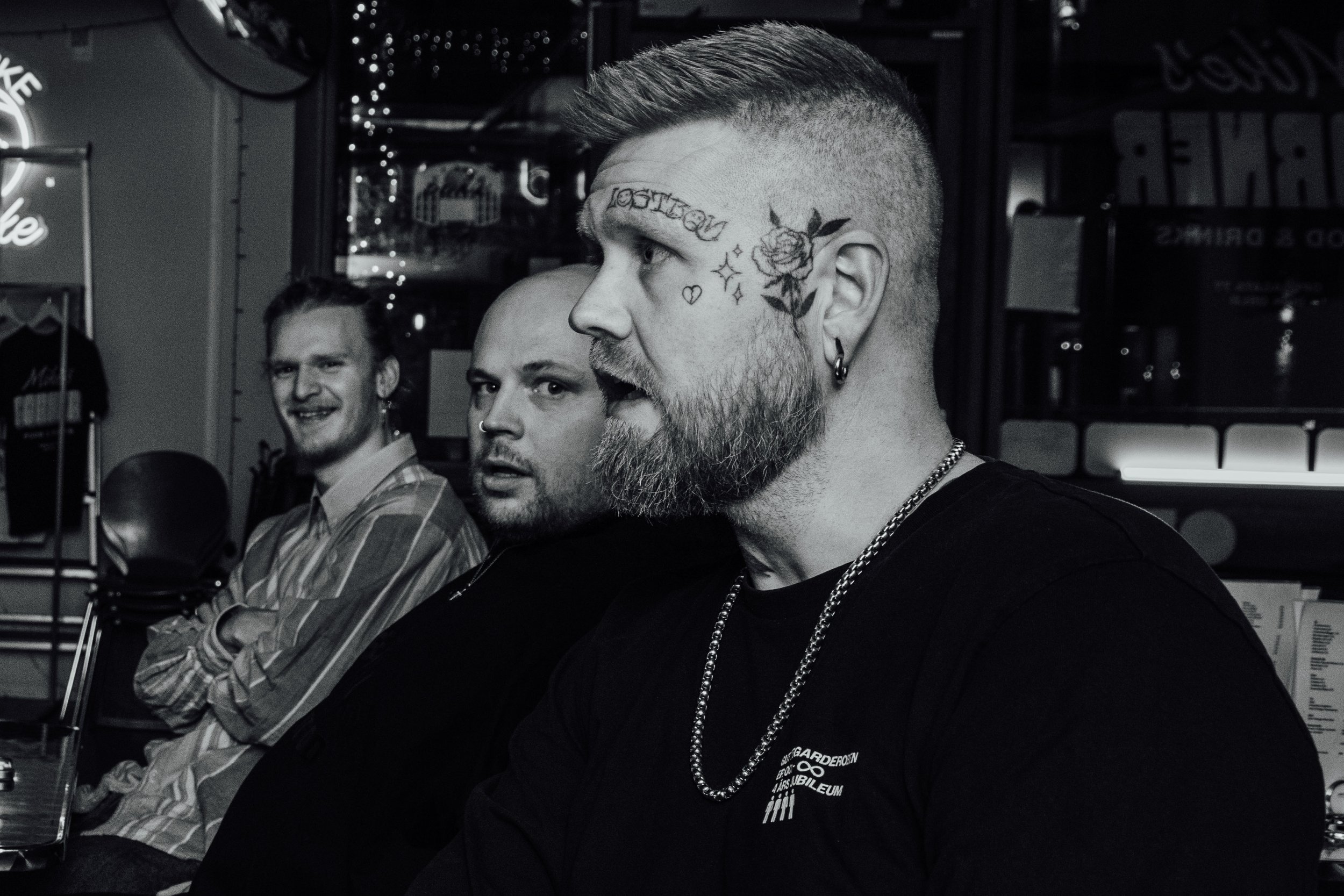 Tre menn sitter på en bar, to av dem ser mot kamera, og den tredje ser ut til å snakke. Den mannlige personen i forgrunn har tatoveringer i ansiktet og på hodet, og har på seg kjede og svart t-skjorte. Skullet speedo lansering
