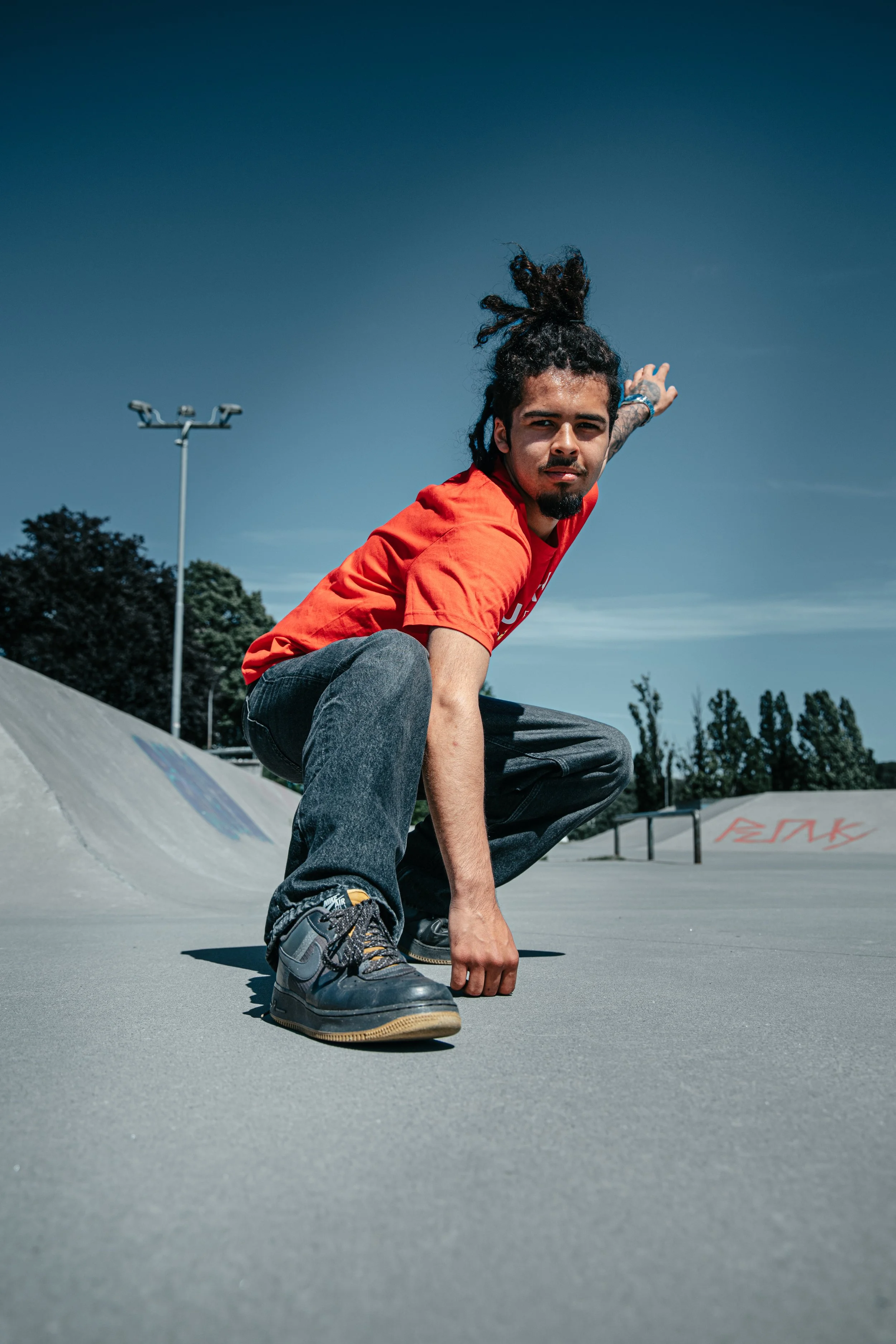 En skateboarder med dreadlocks som bølger i vinden, iført rødt T-skjorte og jeans, i en skatepark under en klar blå himmel. Skate park i Sandefjord
