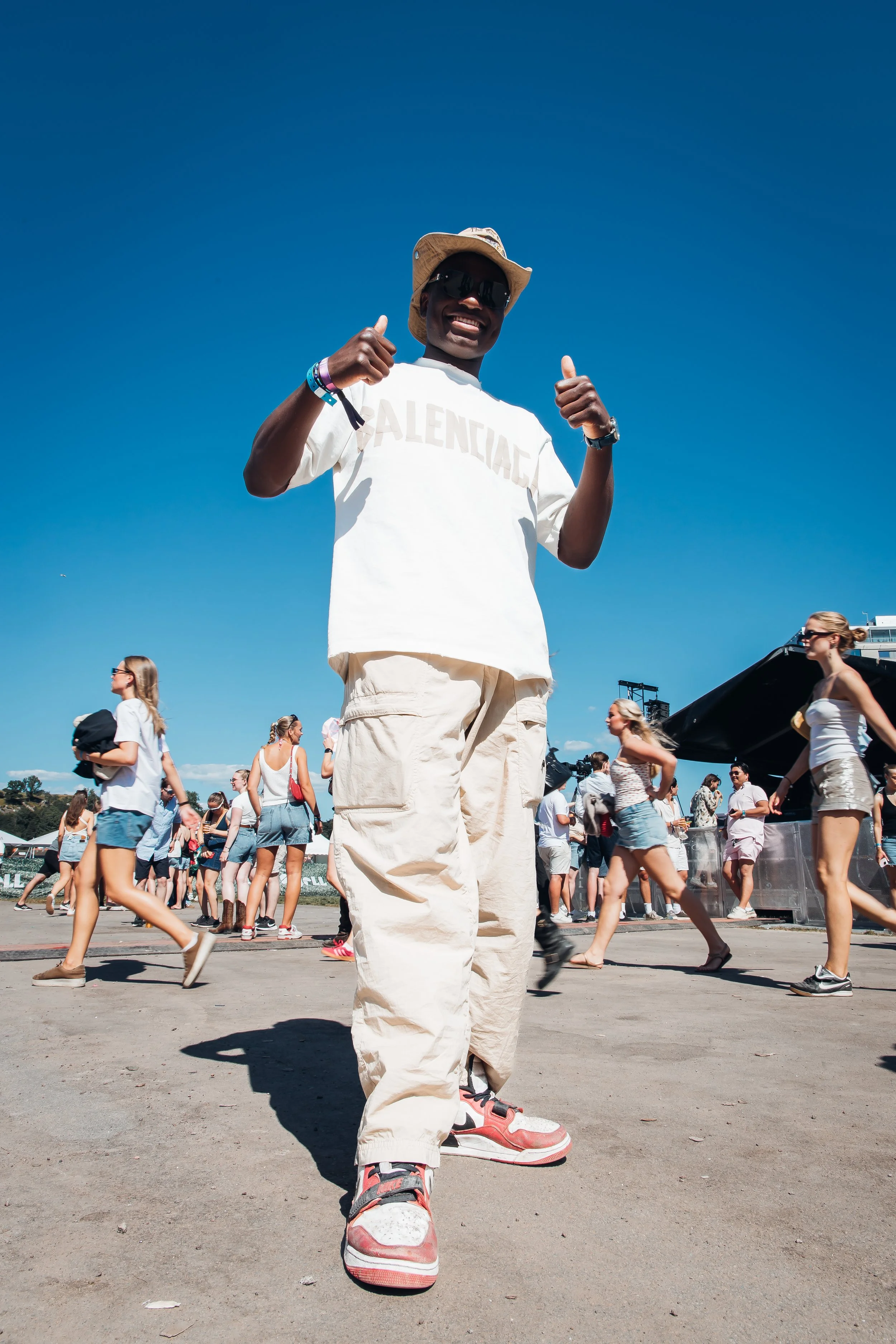 En person står i forgrunnen av et utendørs musikkarrangement, iført hvit t-skjorte med teksten 'BALENCIAGA', beige cargo bukser, fargerike sneakers, en bredtinans-hatt og solbriller, og er omgitt av andre mennesker i sommerklær. Slottsfjell festival