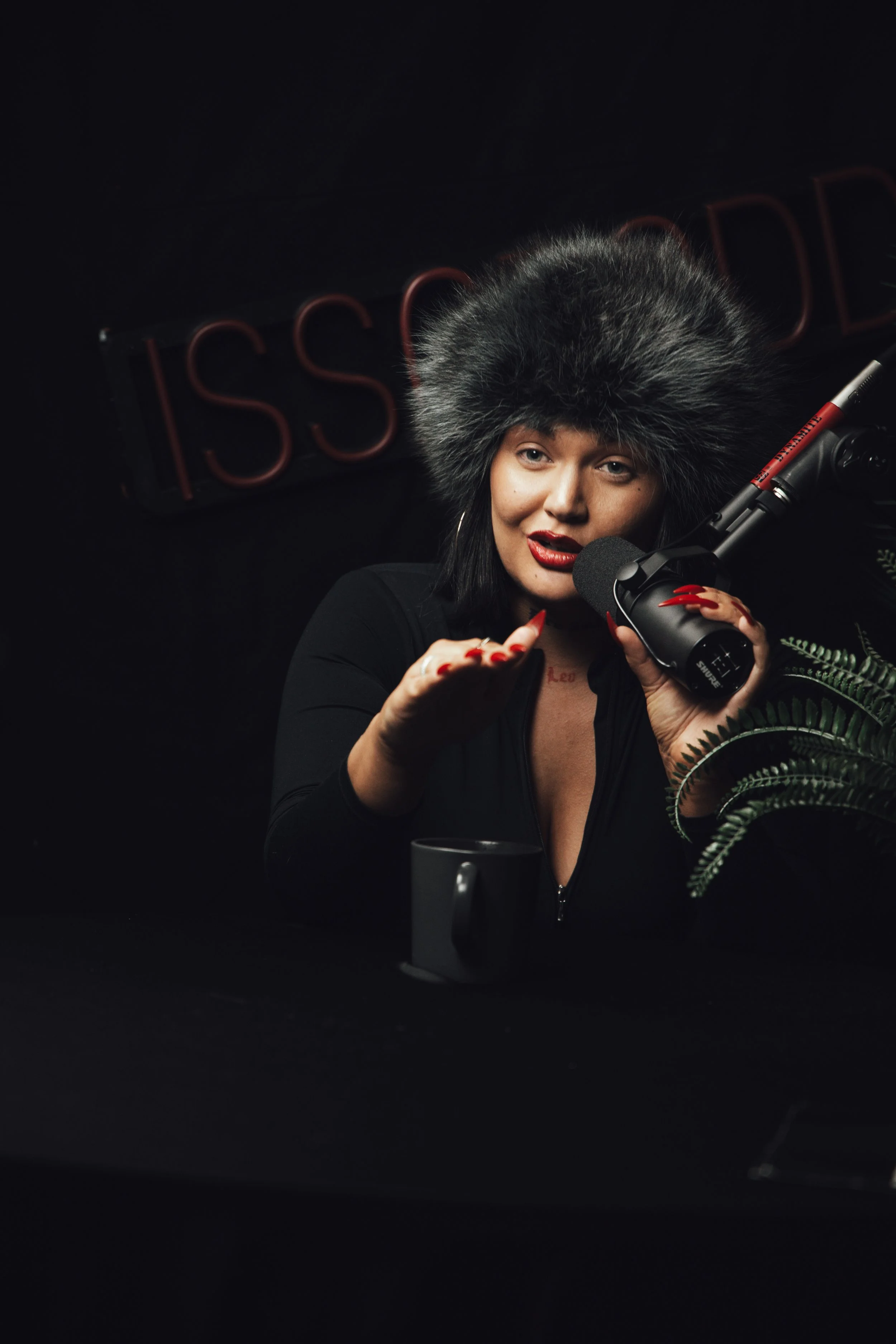 Kvinne med stor pelslue snakker inn i mikrofon, sitter foran en svart bakgrunn med serielt navn og keramikkokopp foran.