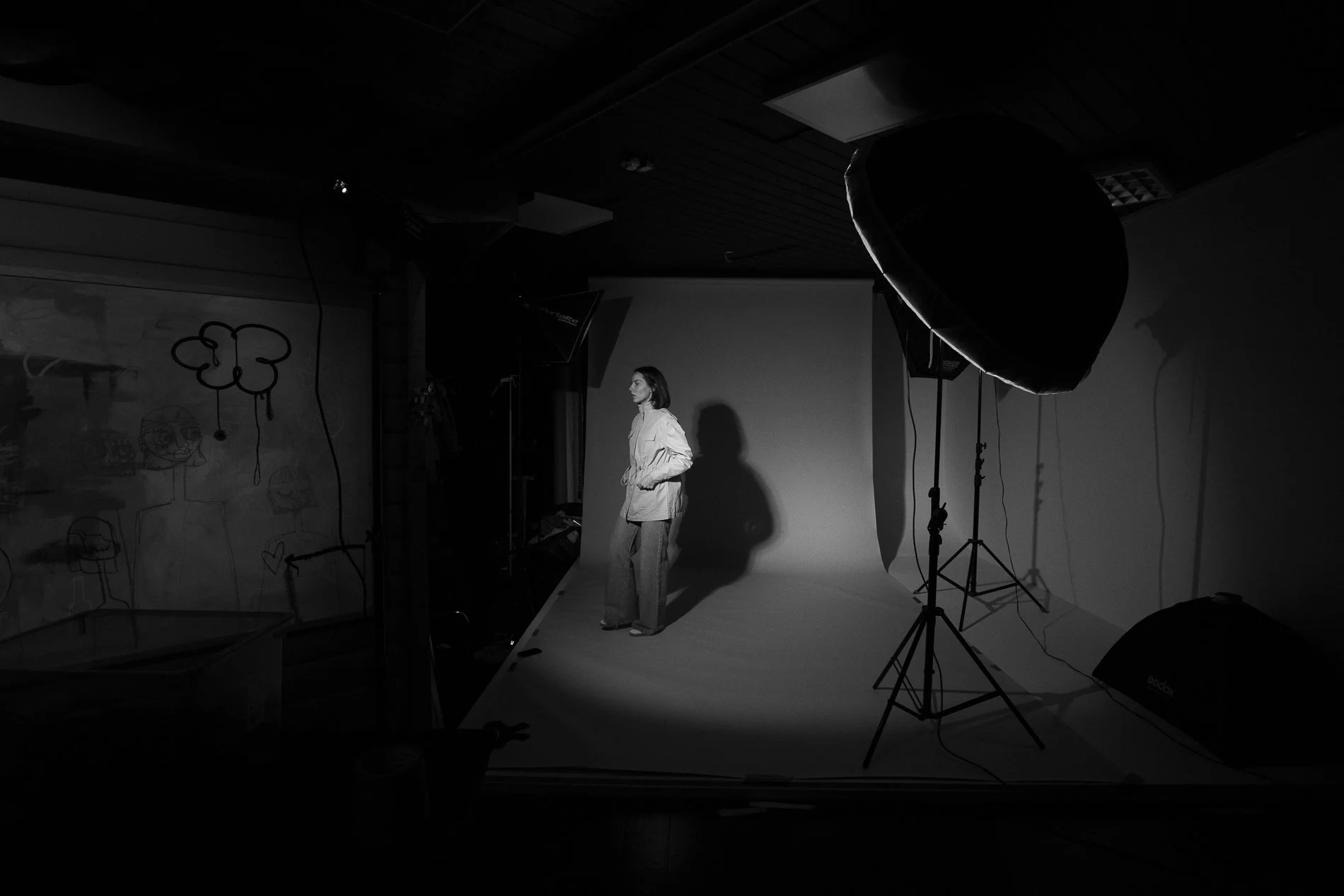 A woman standing on a photography studio set with a plain backdrop, surrounded by lighting equipment, in black and white.