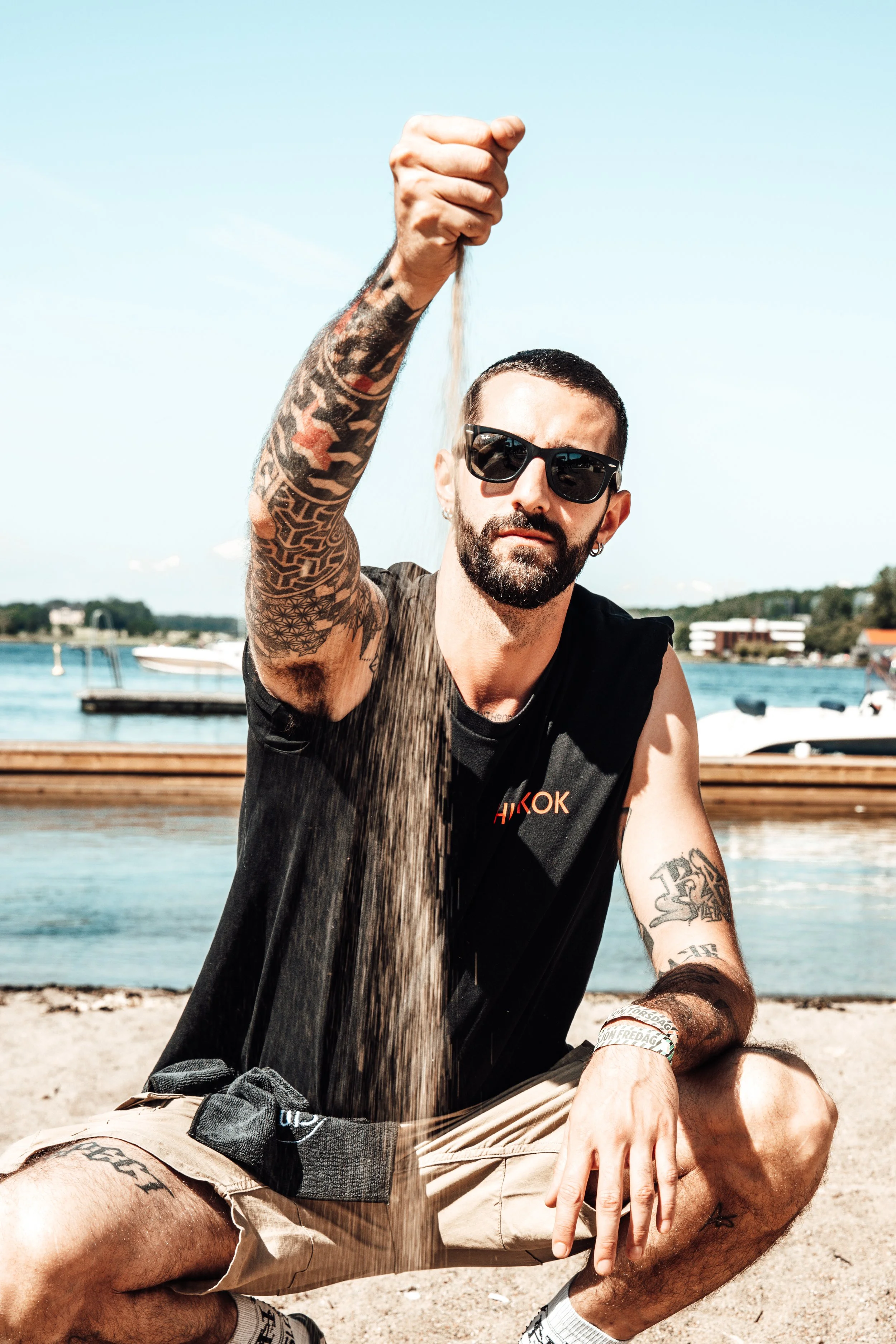 A man with tattoos and sunglasses crouches on a beach, pouring and spilling sand onto the ground with a stern expression, near water and boats in the background.