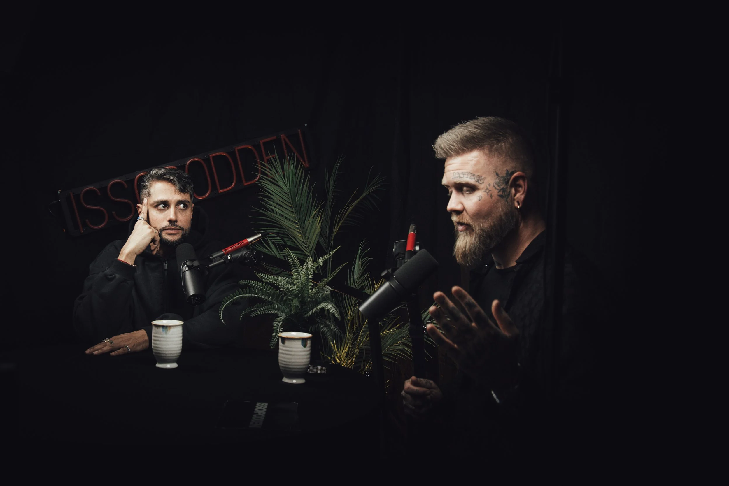 To menn snakker i mikrofoner under en podkast-innspilling, med planter og et svart bakteppe, der en av mennene har tatoveringer og skjegg.