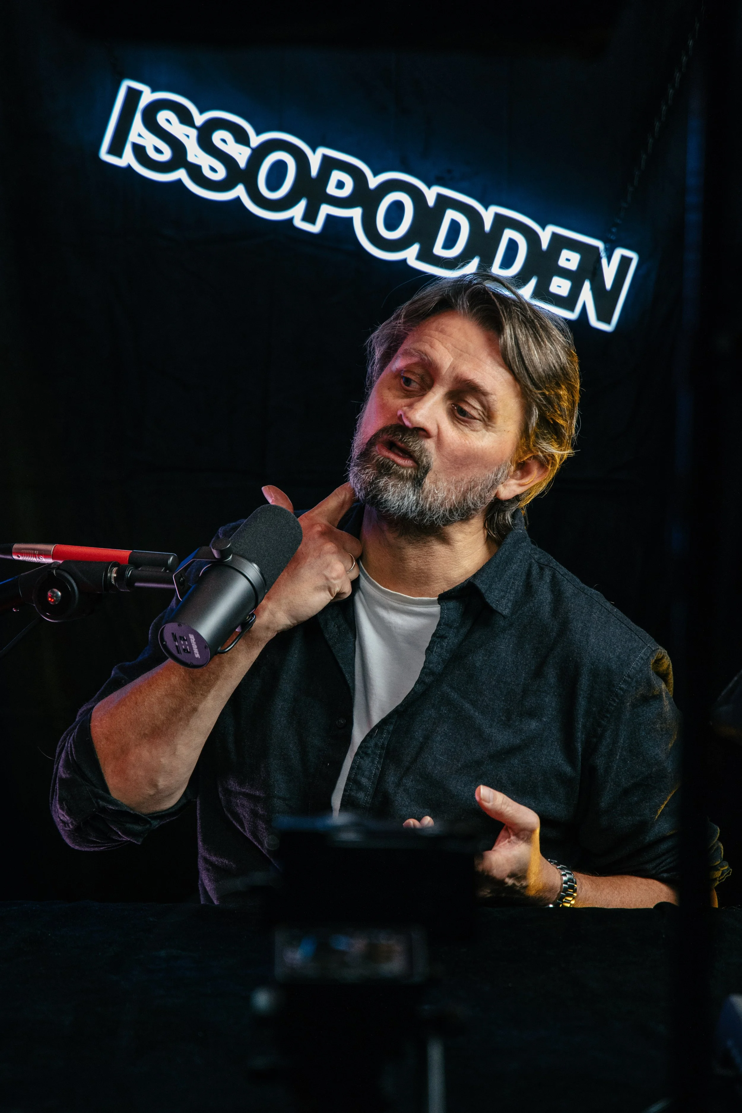 En mann med grått skjegg og hår som sitter foran en mikrofon og peker på nakken sin, med en neonlysknekt som sier 'ISSOPODDEN' i bakgrunnen. Thomas Numme