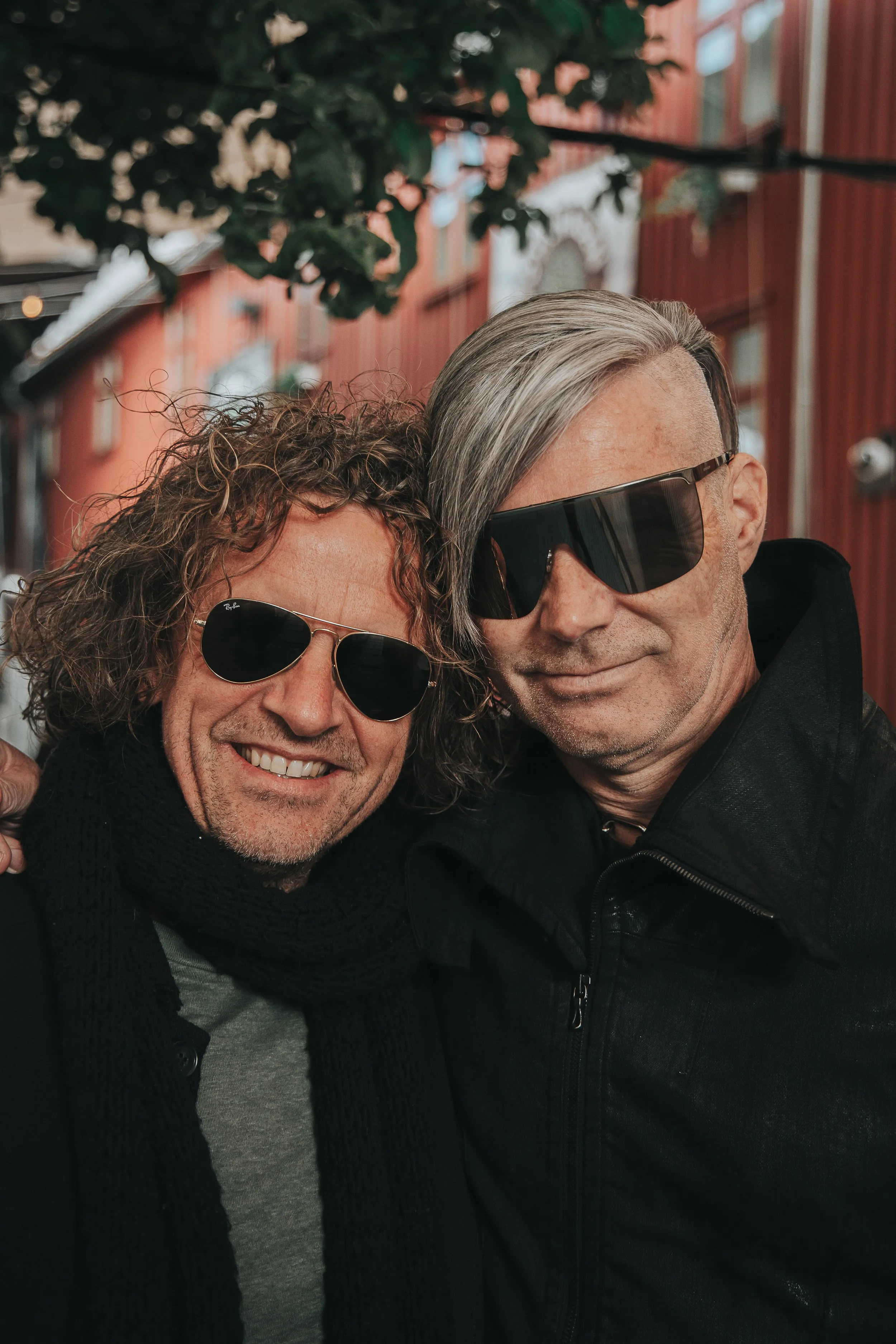 To menn med solbriller som smiler og tar selfie sammen utenfor en bygning med rødt utseende. Seigmen 