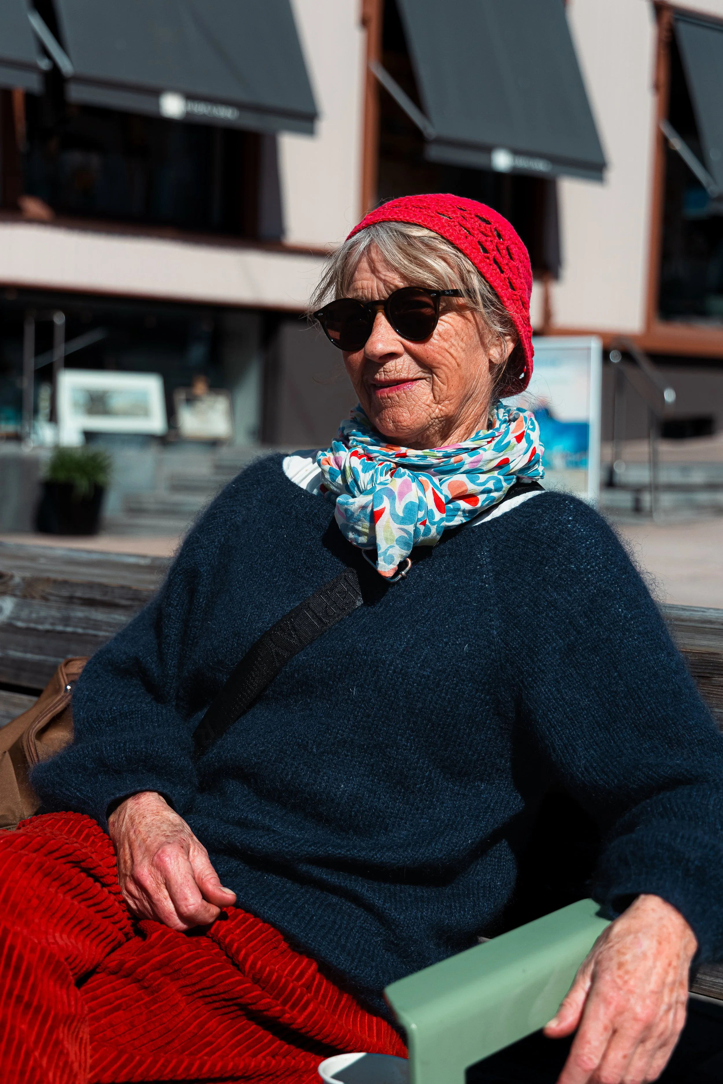 En eldre kvinne med solbriller, rød sylkofte og skjerf, sitter utendørs i solen med en bygning i bakgrunnen.
